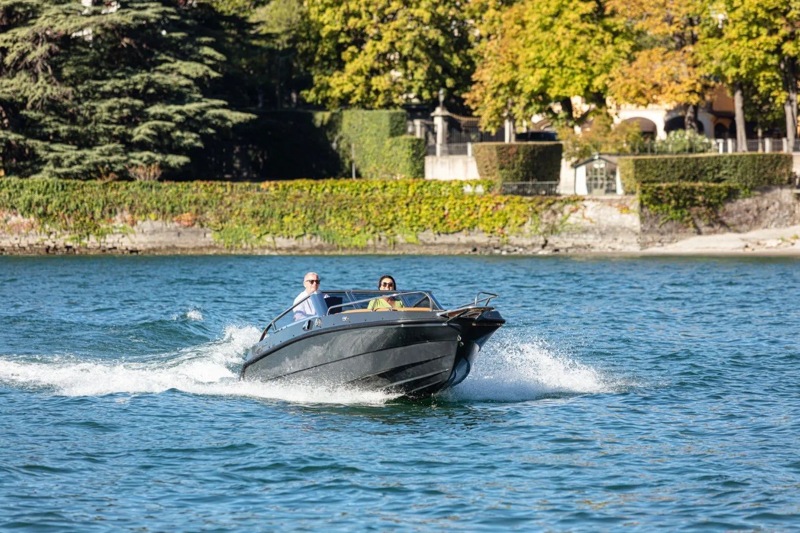 Two people in black hull boat on a plane going through water passing houses green foliage in the background