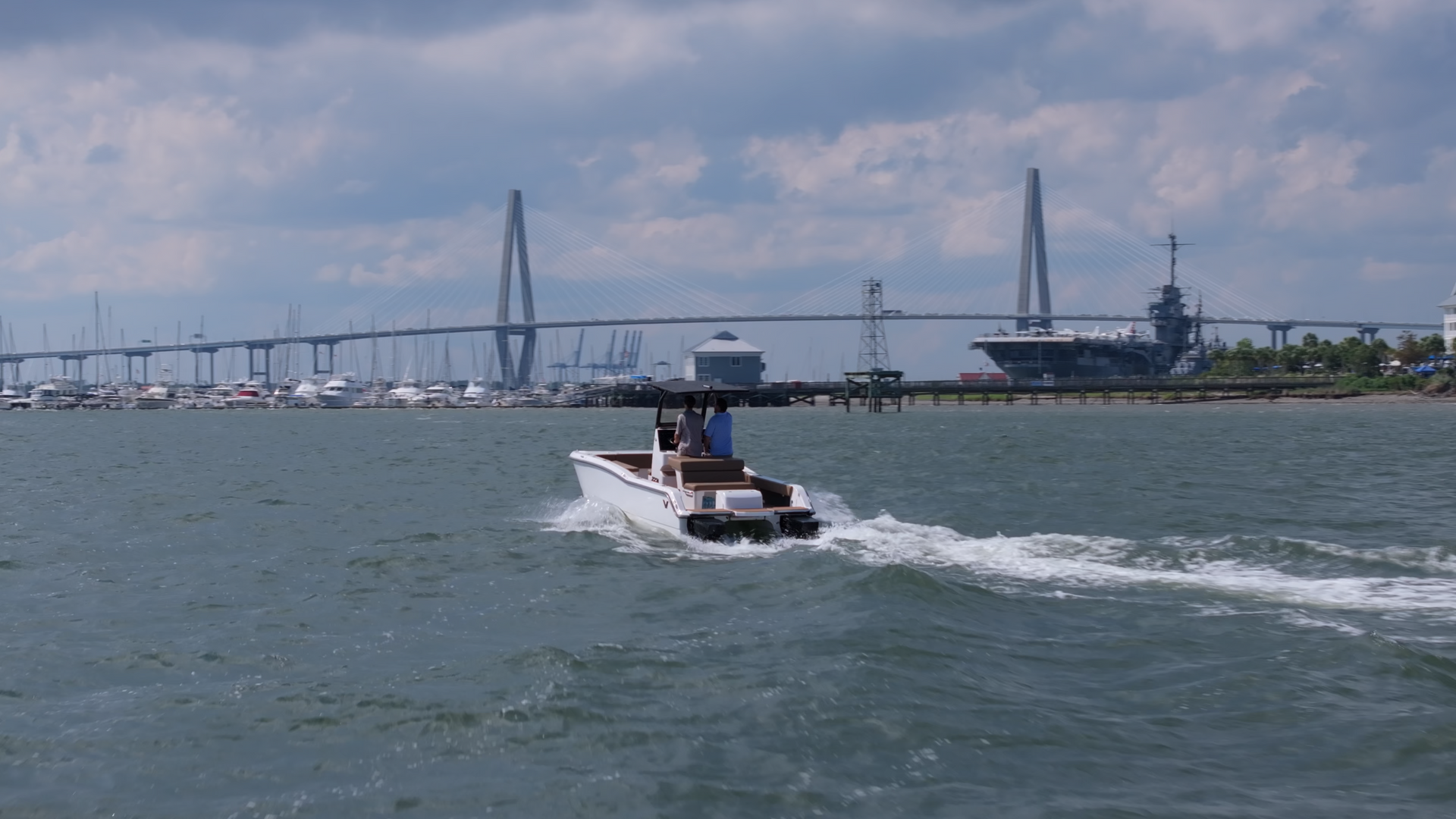 White T-Top boat with twin outboard motors cruising towards a bridge in water shore in site. In background other bigger boats and ships