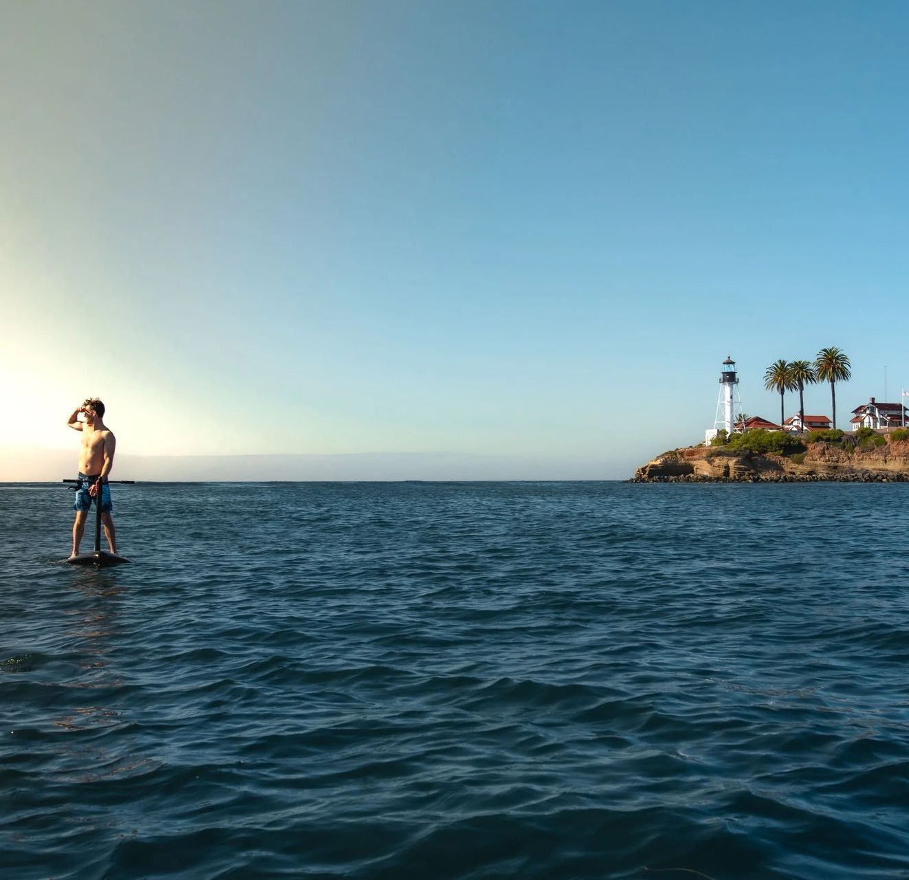 A man is standing on a paddle board in the ocean with a lighthouse in the background.