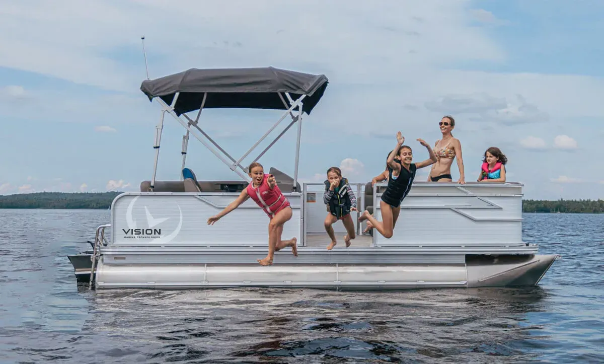 four kids and a woman on a light grey pontoon. Three kids are jumping off boat into water with life jackets on. There is a dark grey canopy above the captain seat towards the back of the boat.