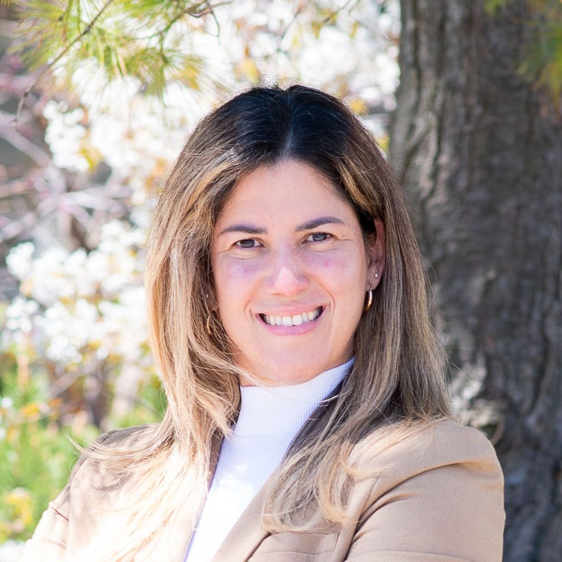 Smiling person with long hair in a beige coat outdoors, standing near trees and sunlight