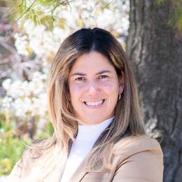 Smiling woman with long brown hair wearing a beige coat outdoors by trees