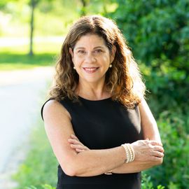 Smiling woman in a black sleeveless top stands outdoors with arms crossed on a sunlit path.