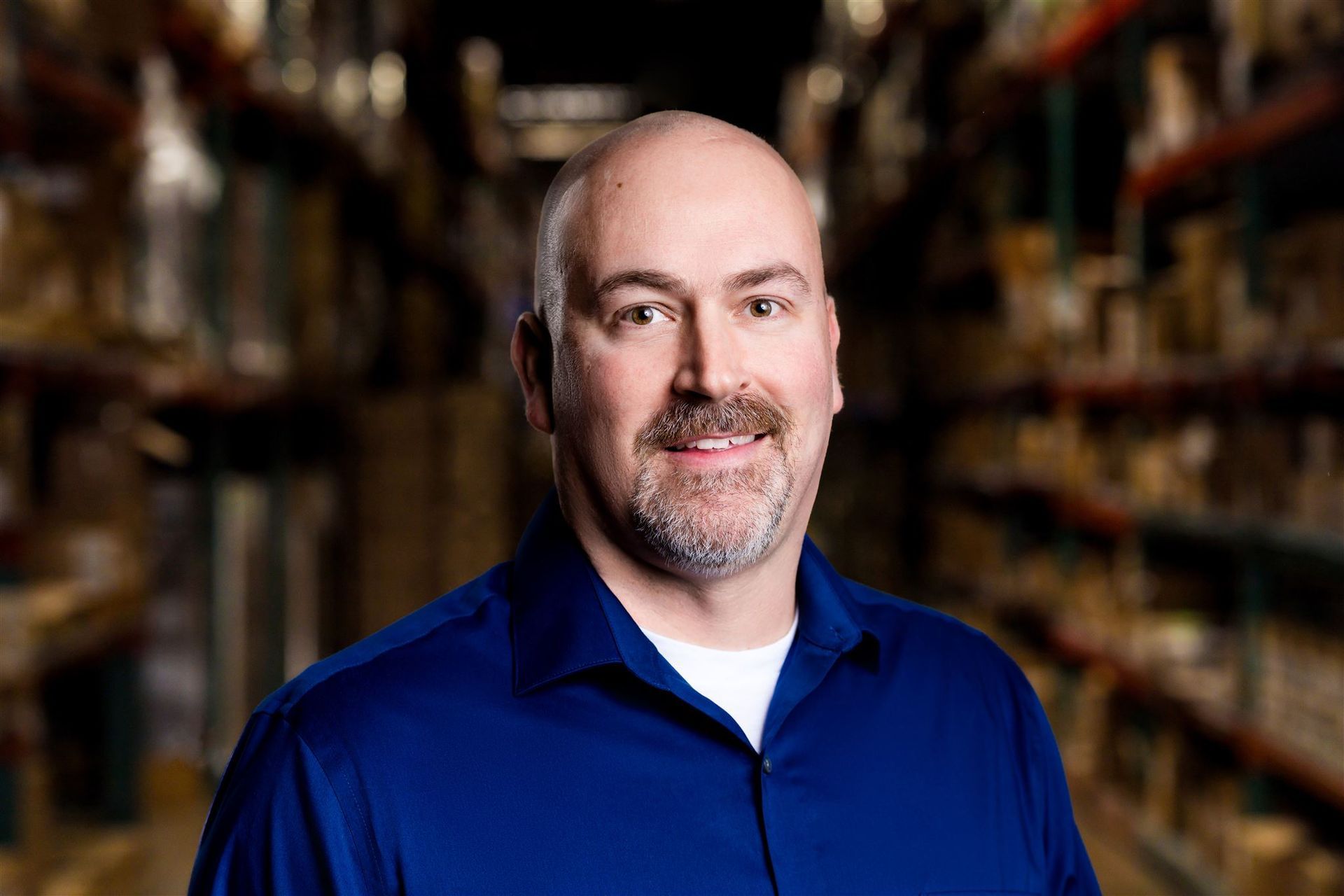 Smiling man in a blue shirt standing in a warehouse aisle with shelves in the background