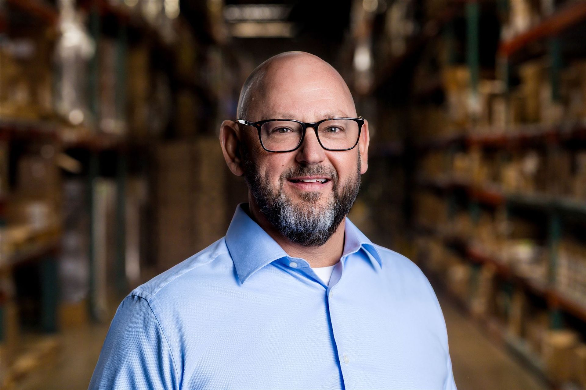 Smiling bald man with glasses in a light blue shirt, standing in a blurred warehouse aisle