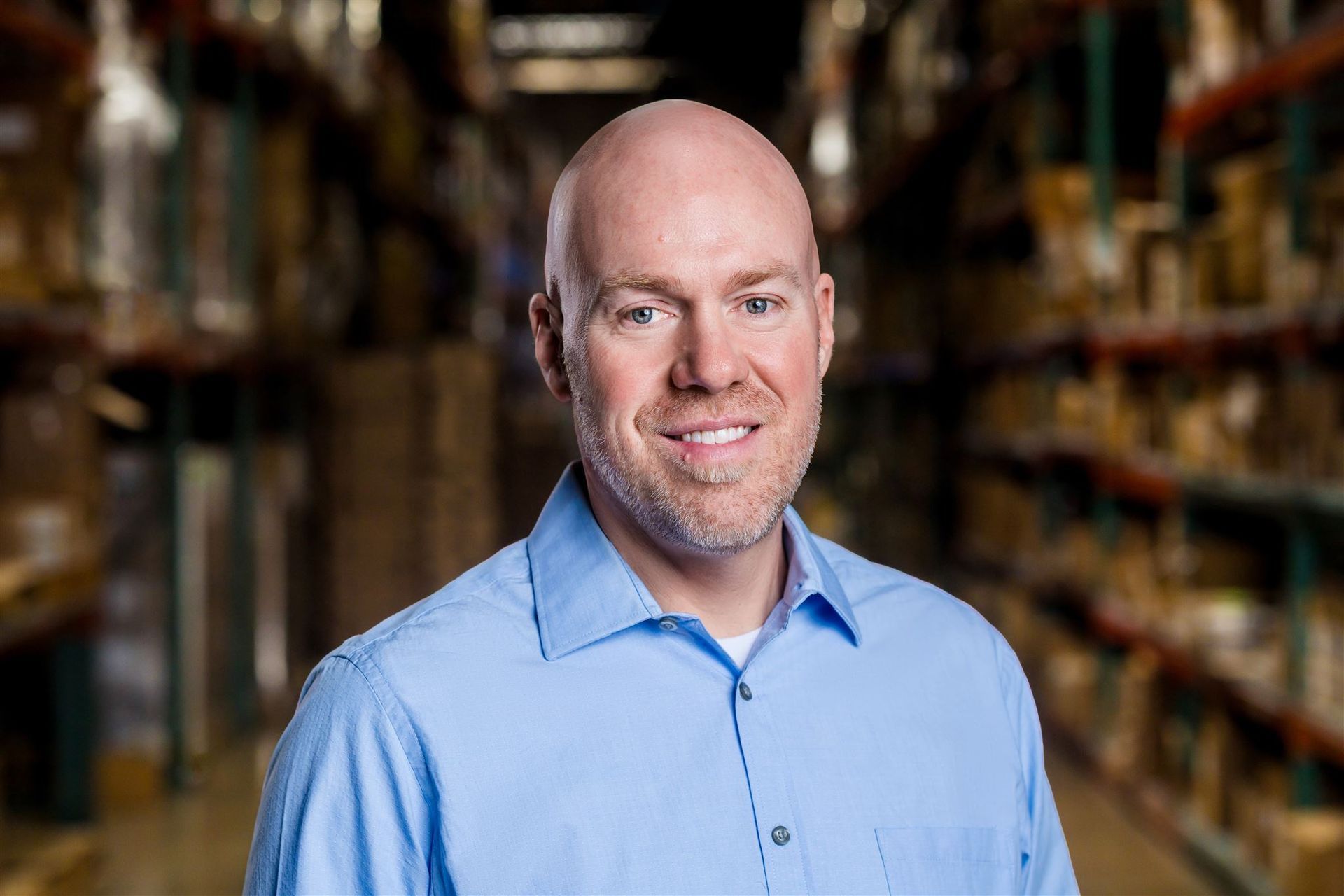 Man in a light blue shirt smiling in a warehouse aisle with shelves in the background