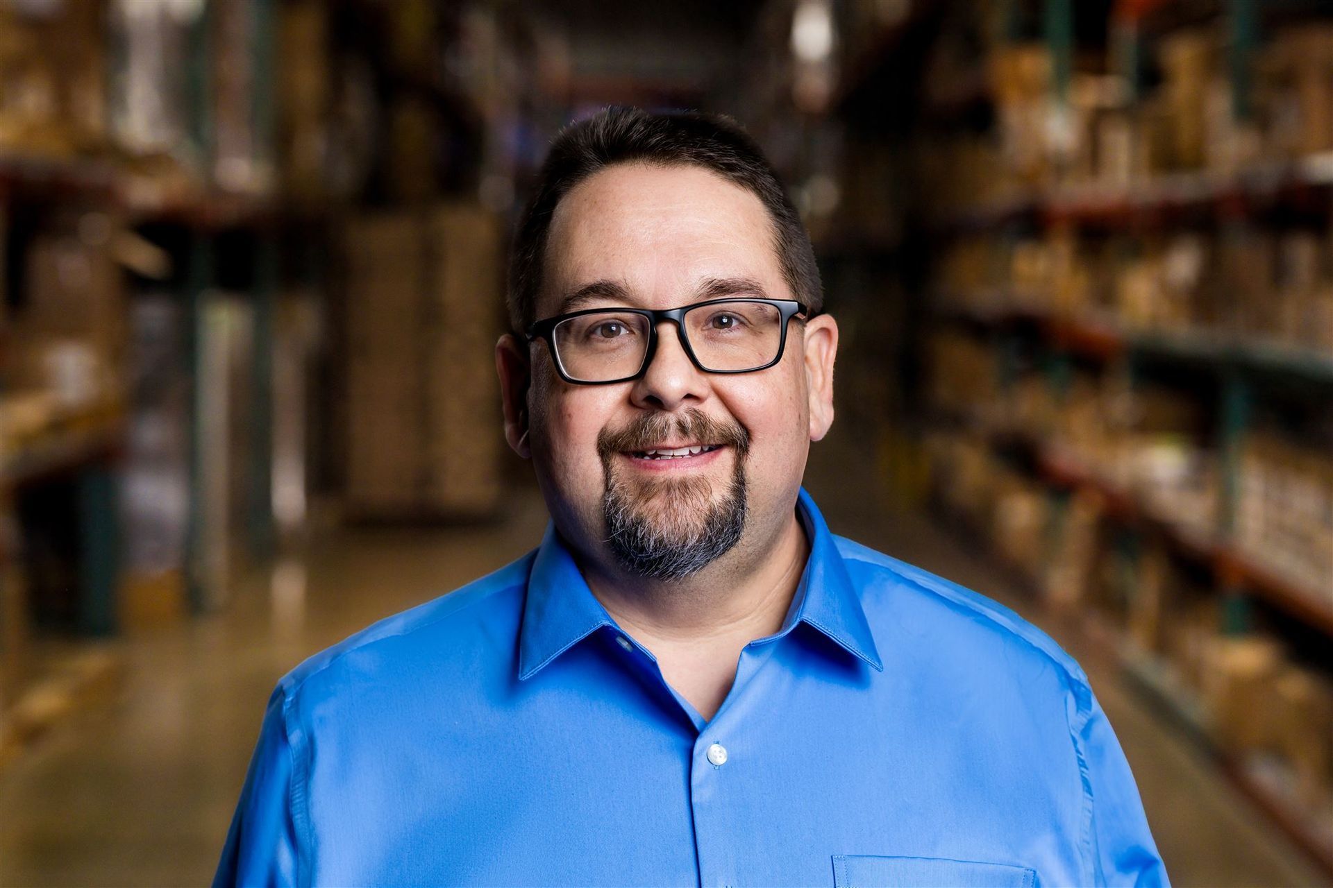 Smiling man in a blue shirt standing in a store aisle with shelves blurred behind him