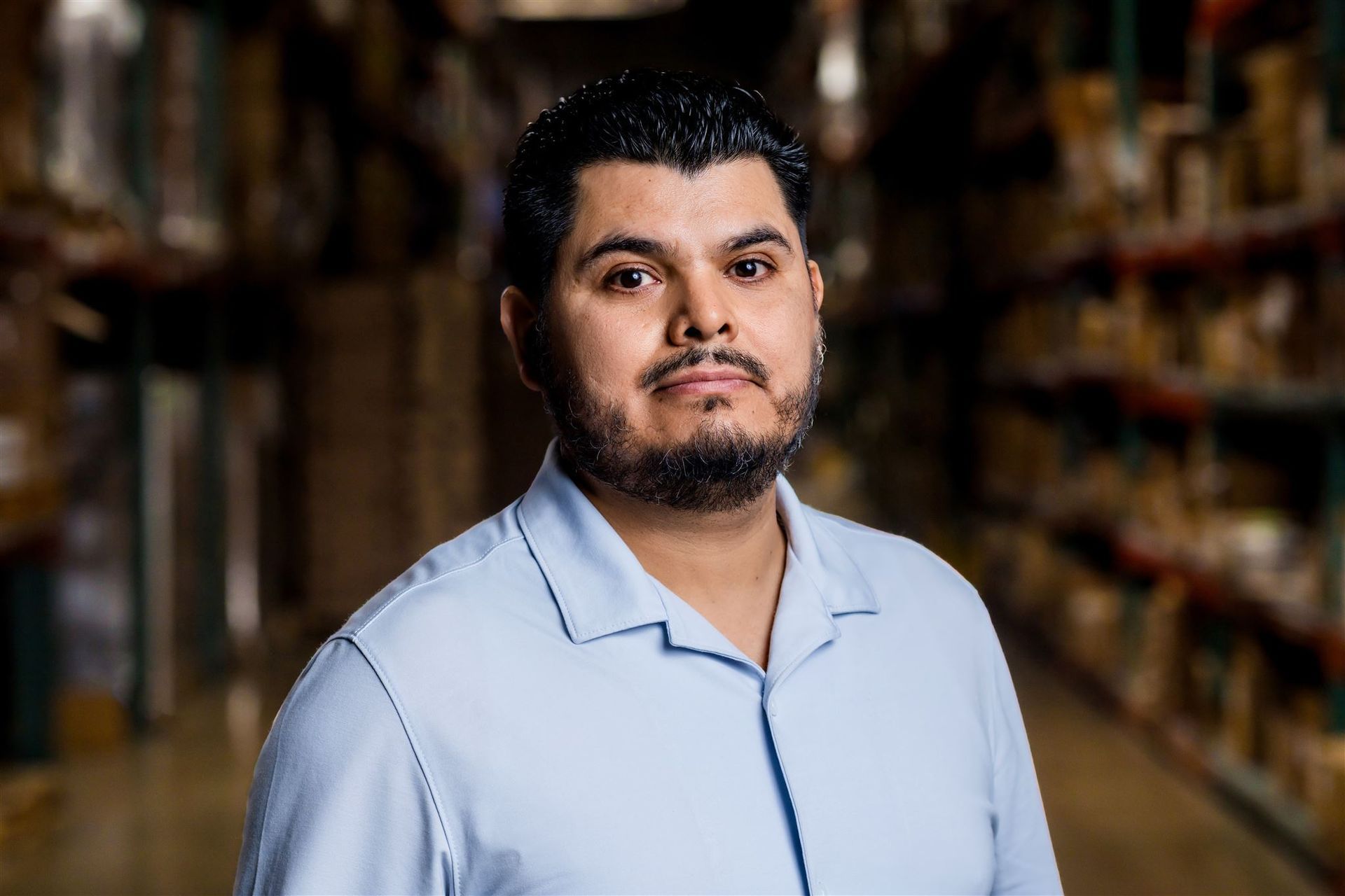 Man standing in a warehouse aisle, wearing a light blue shirt, looking at the camera.