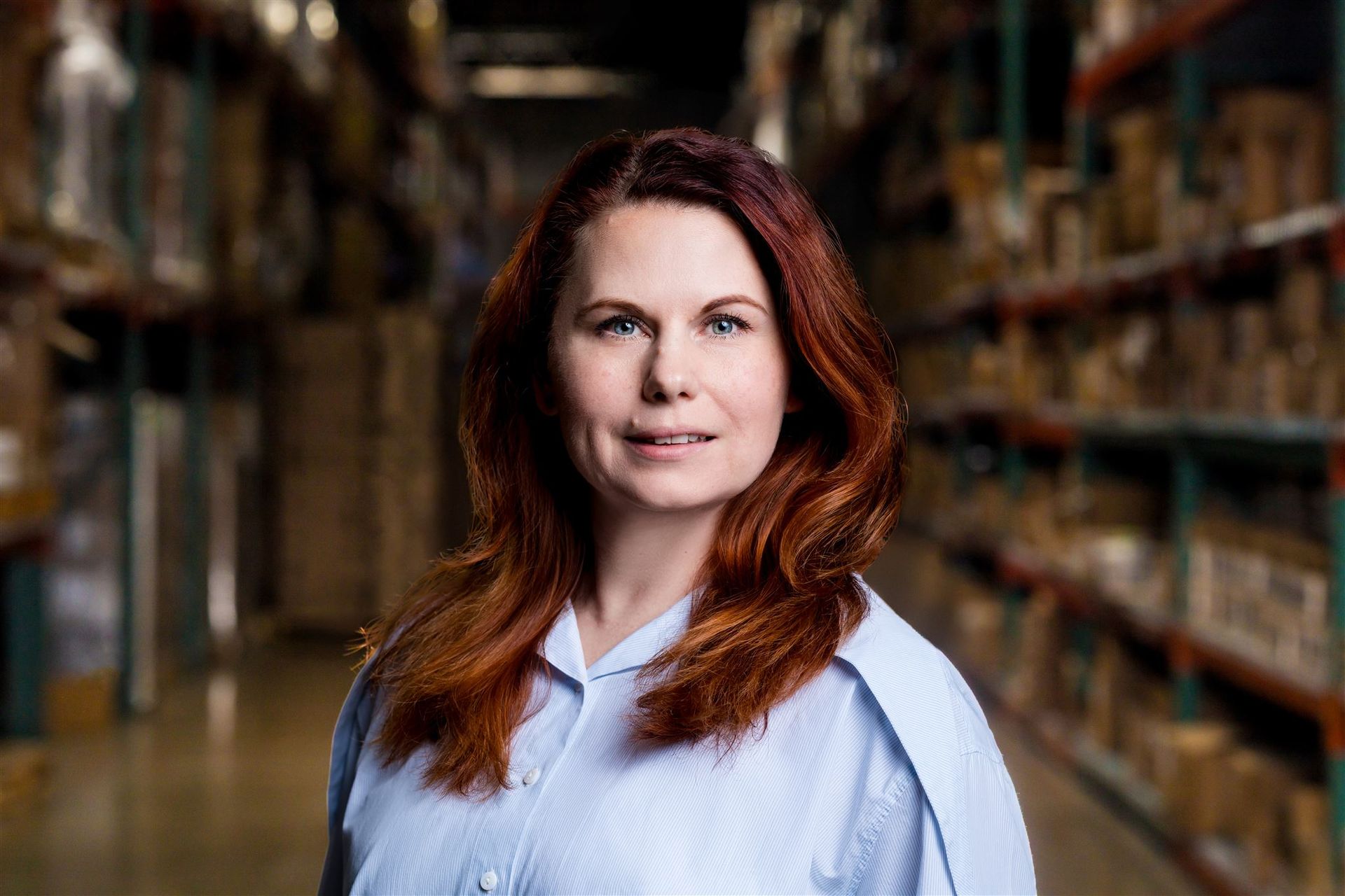 Woman with auburn hair standing in a library aisle, wearing a light blue shirt and looking at the camera
