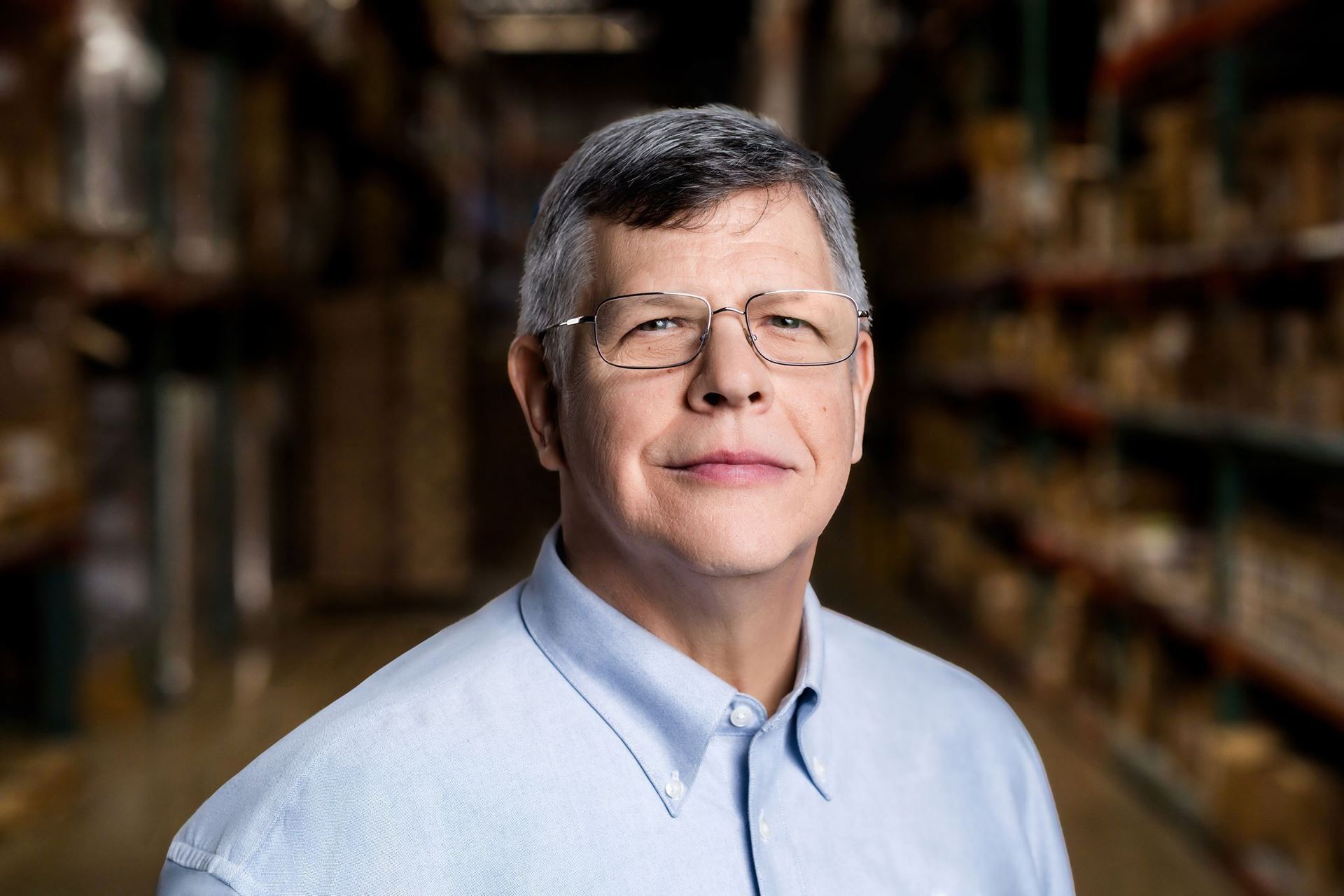Man in a light blue shirt standing in a blurred bookstore aisle, facing the camera with a slight smile
