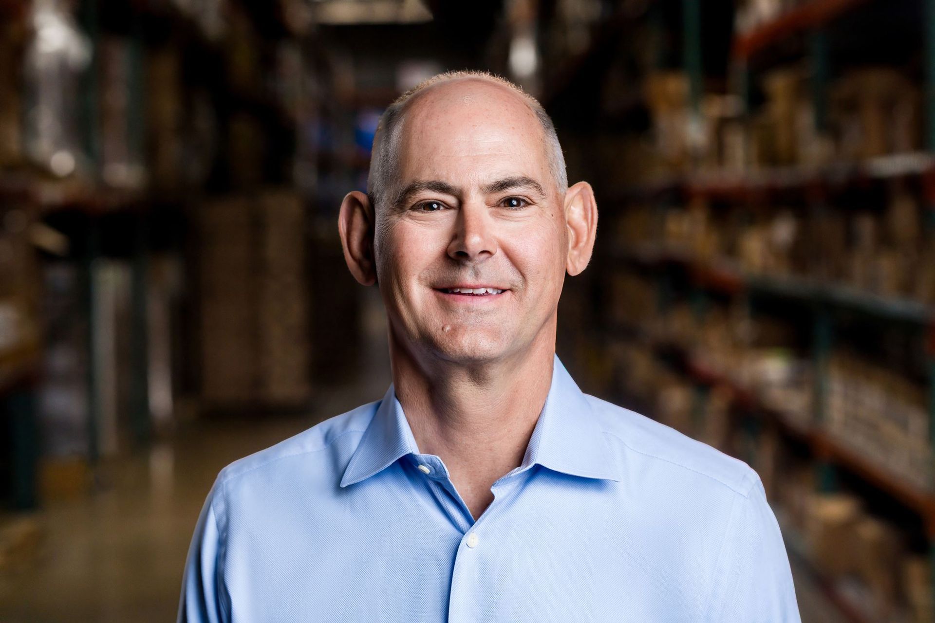 Portrait of a smiling bald man in a light blue shirt, standing in a blurred bookstore aisle