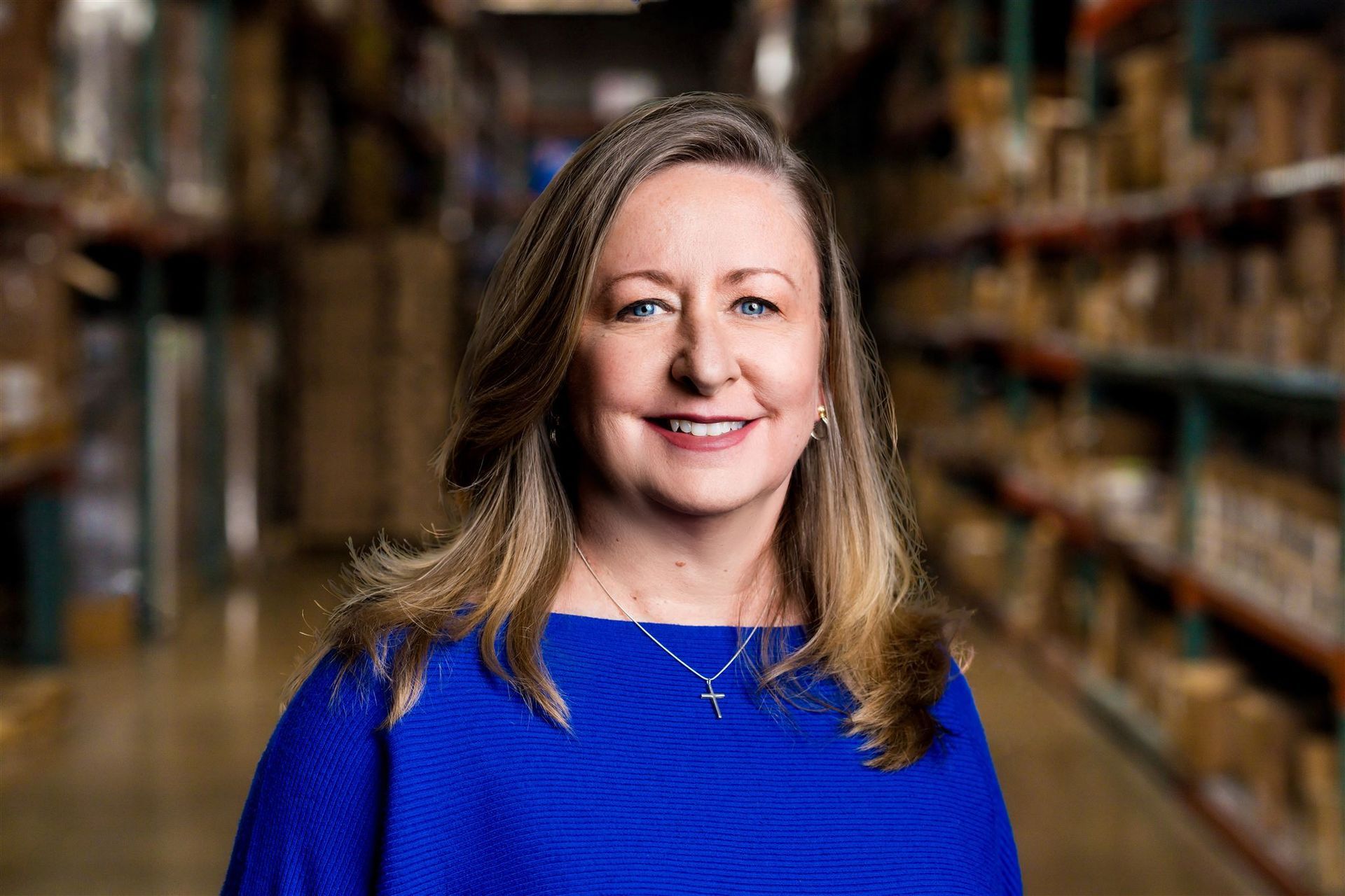 Woman in a blue sweater smiling in a warehouse aisle with shelves in the background
