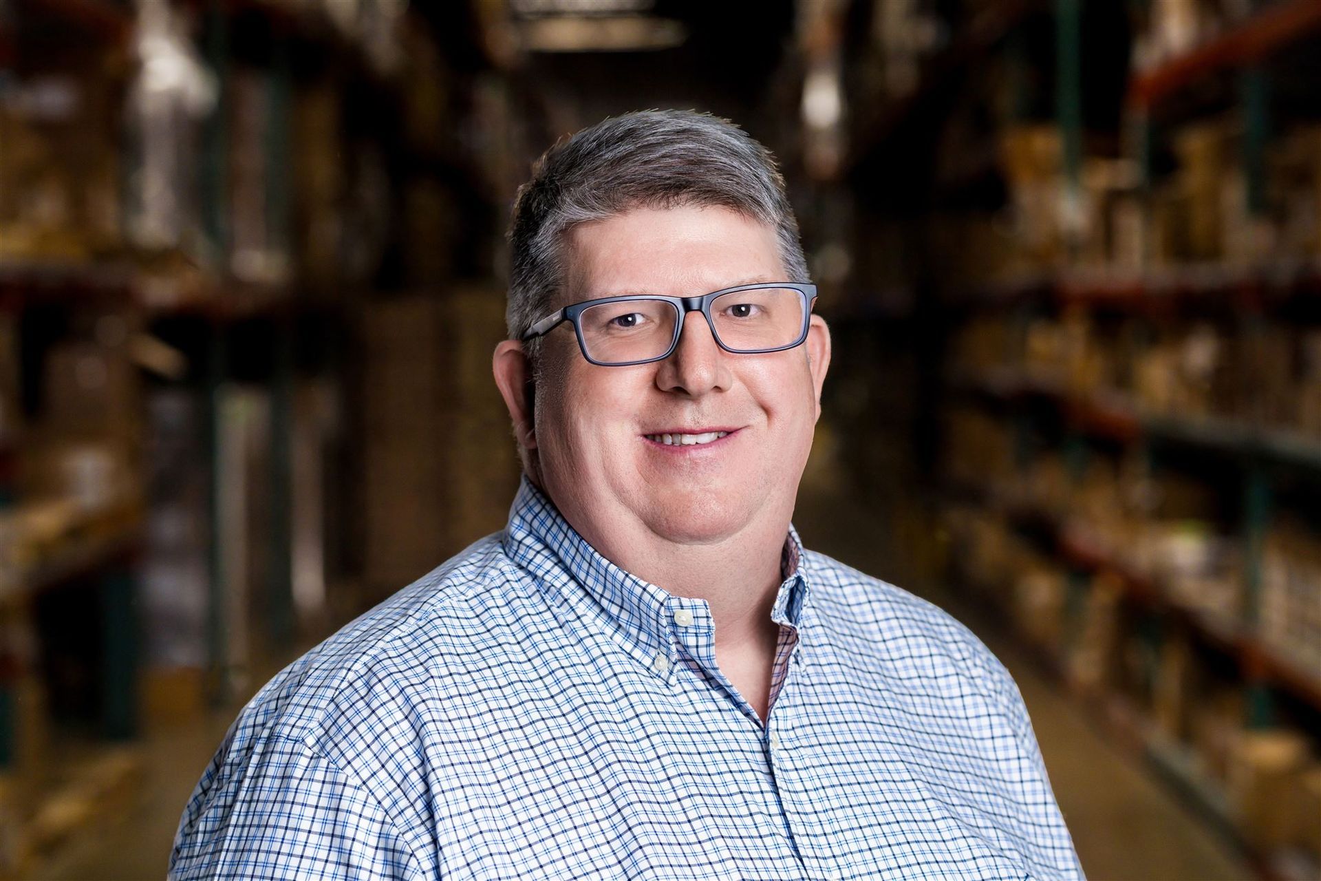 Man standing in a warehouse aisle, smiling, wearing glasses and a blue checkered shirt.