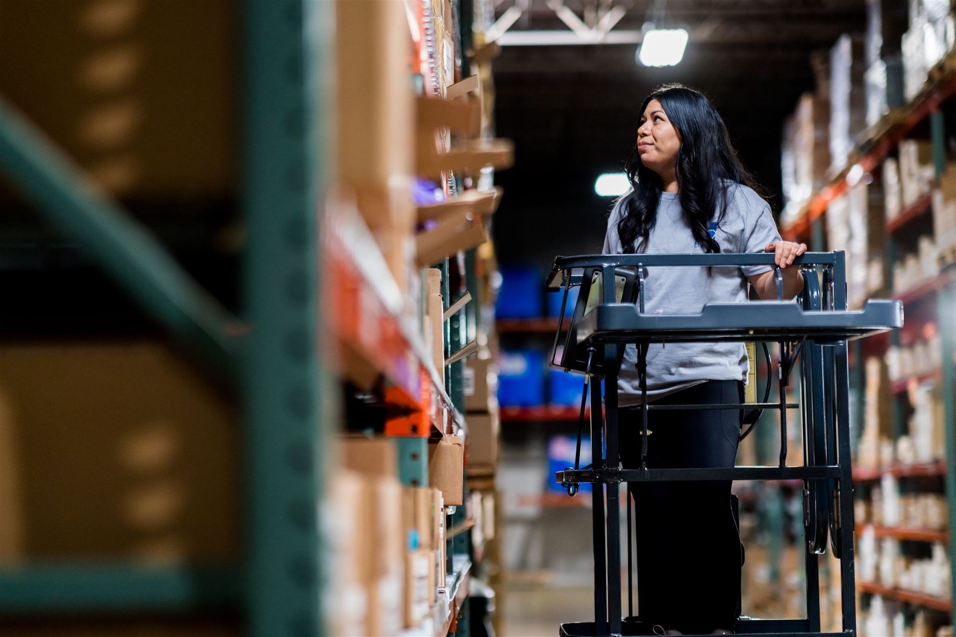 Person standing with a cart in a warehouse aisle, reaching up toward shelves.