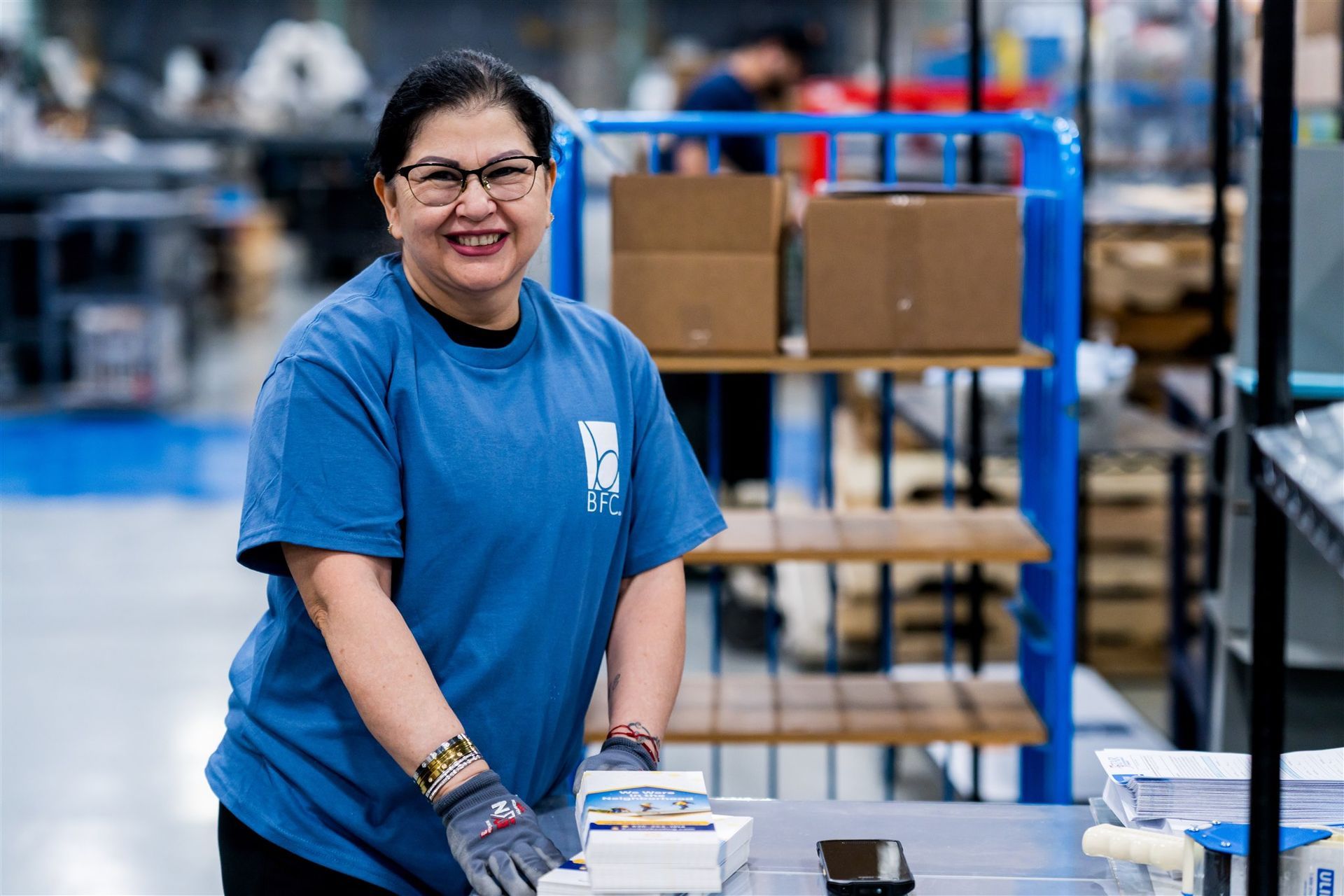Woman in a blue shirt smiling in a warehouse, standing by a table with boxes and shelves in the background