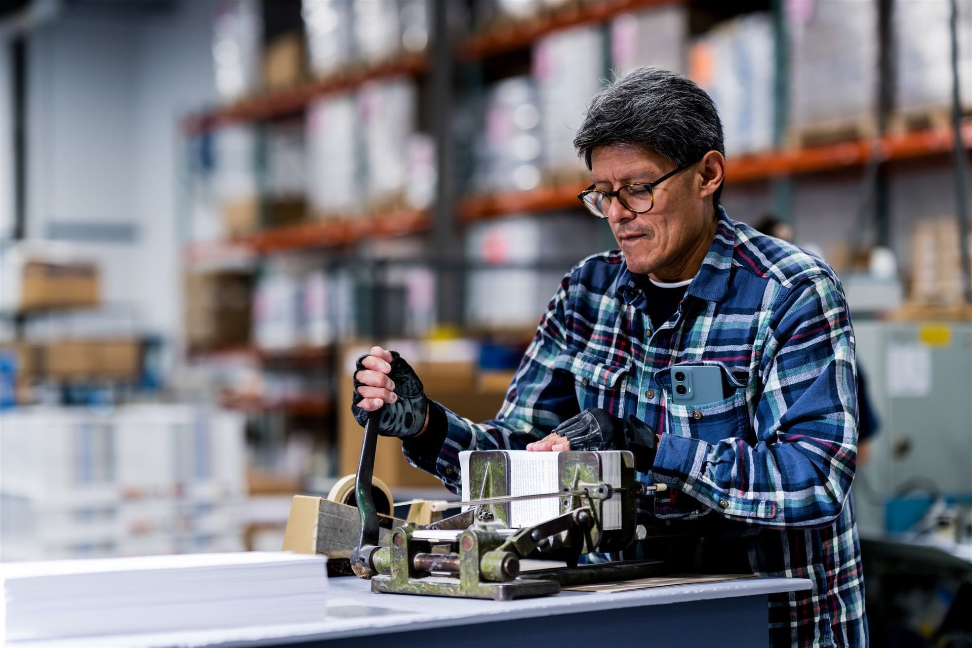 Worker in a blue plaid shirt using a tool on equipment in a warehouse workspace