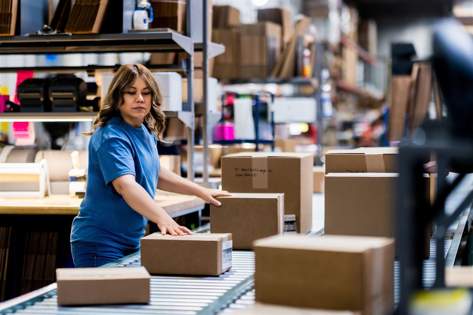Warehouse worker in blue shirt sorting packages on a conveyor belt in a busy storage area