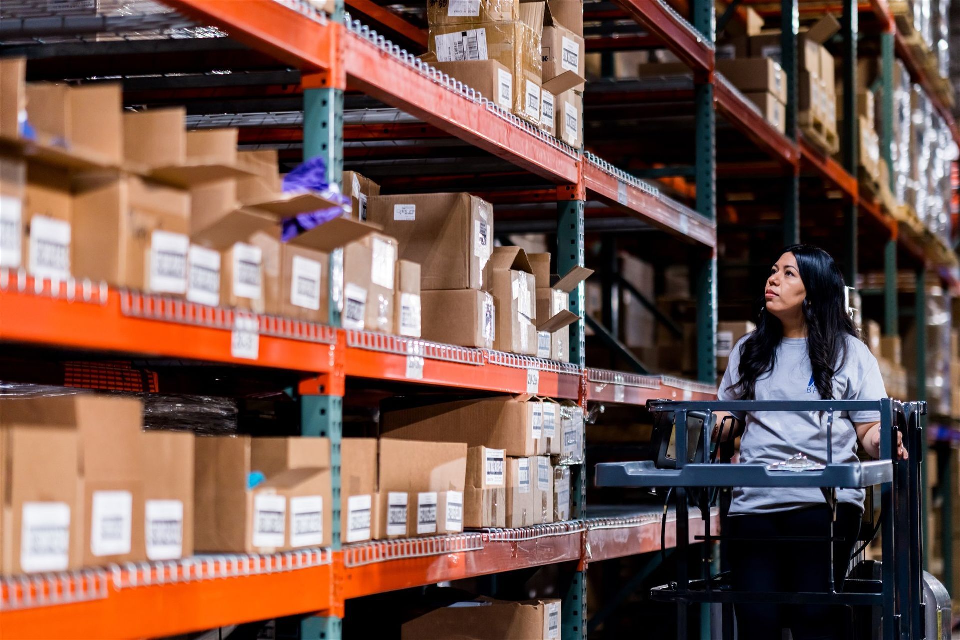 Woman on a warehouse lift reaching toward stacked cardboard boxes on orange shelves