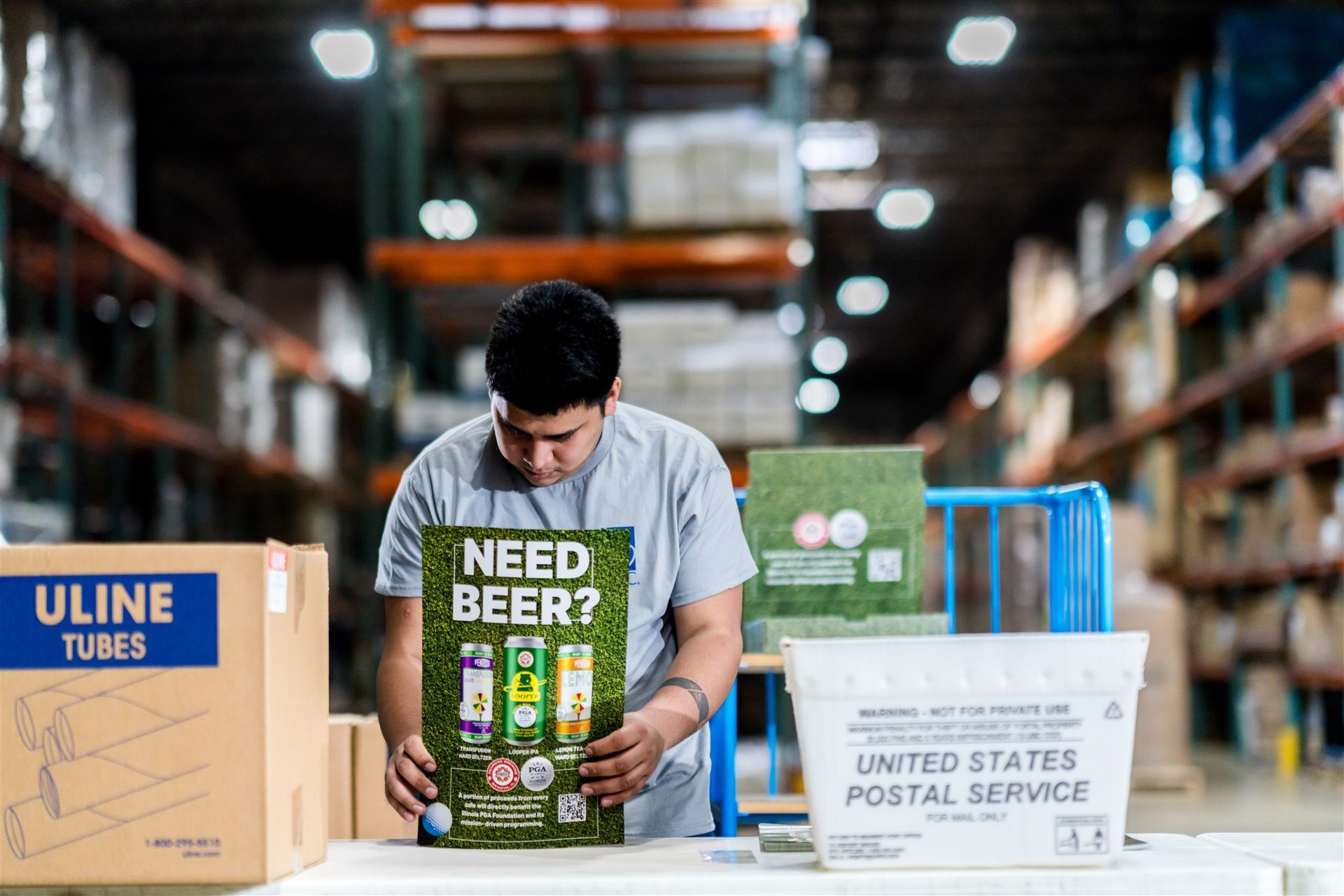 Man packing beer bottles in a warehouse aisle beside Uline and USPS boxes