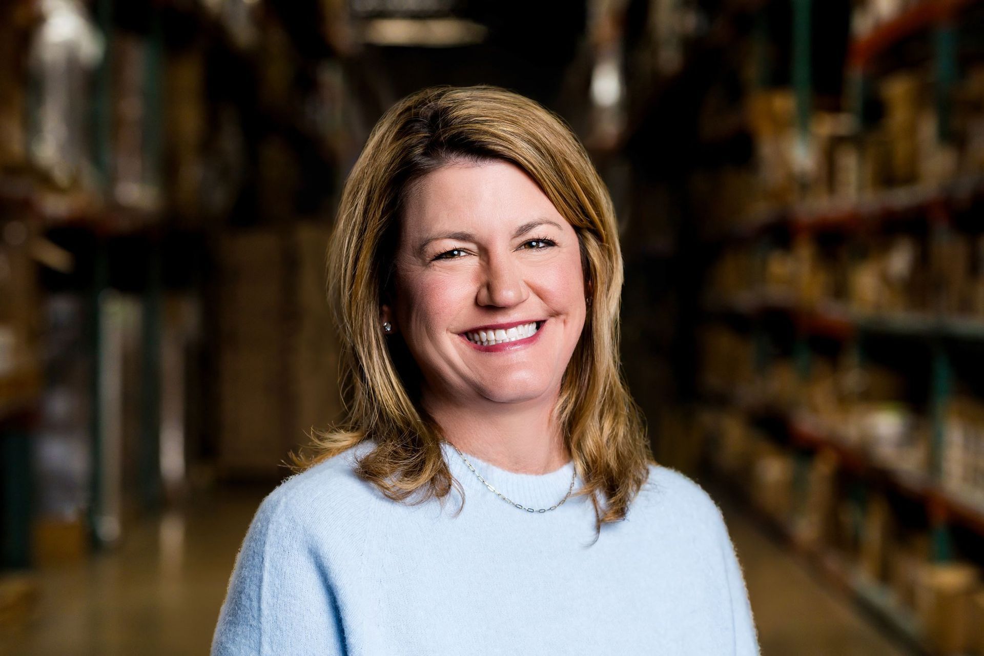 Smiling man in a light blue shirt standing in a warehouse aisle with shelves in the background