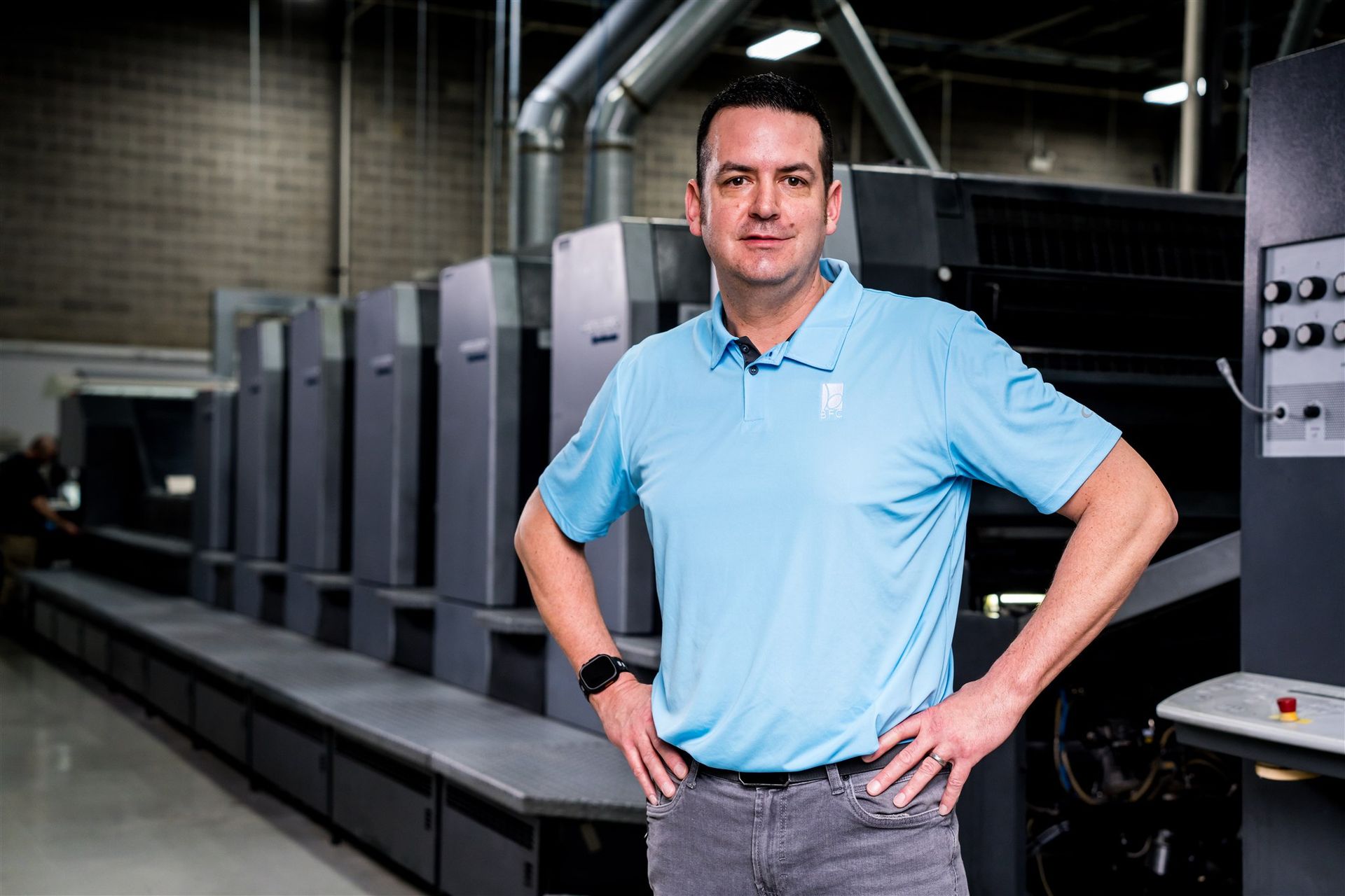 Man standing in a factory beside large industrial printing equipment, wearing a light blue polo shirt.