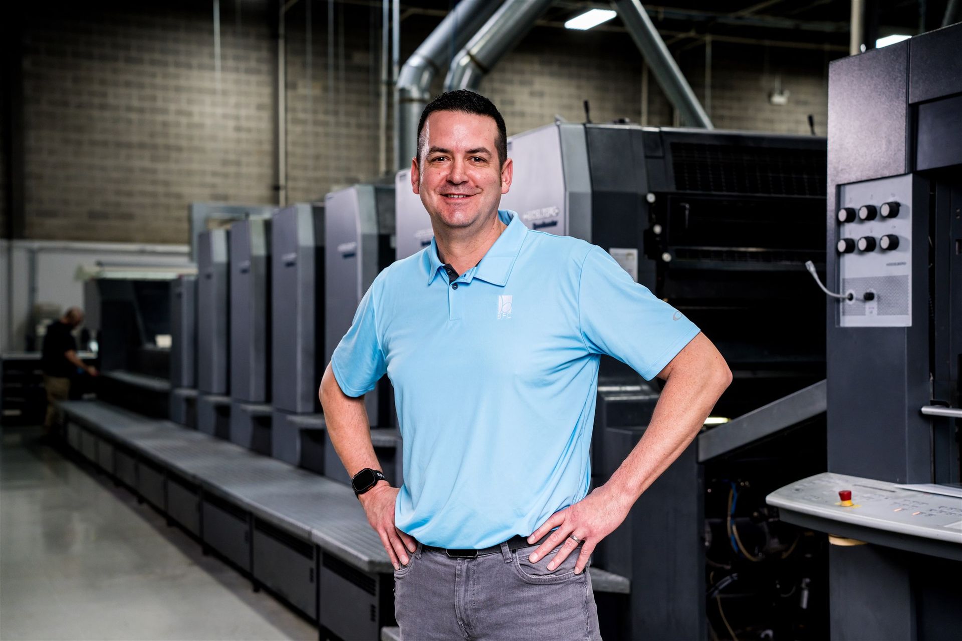 Man in a light blue polo stands beside a large industrial printing press in a factory.