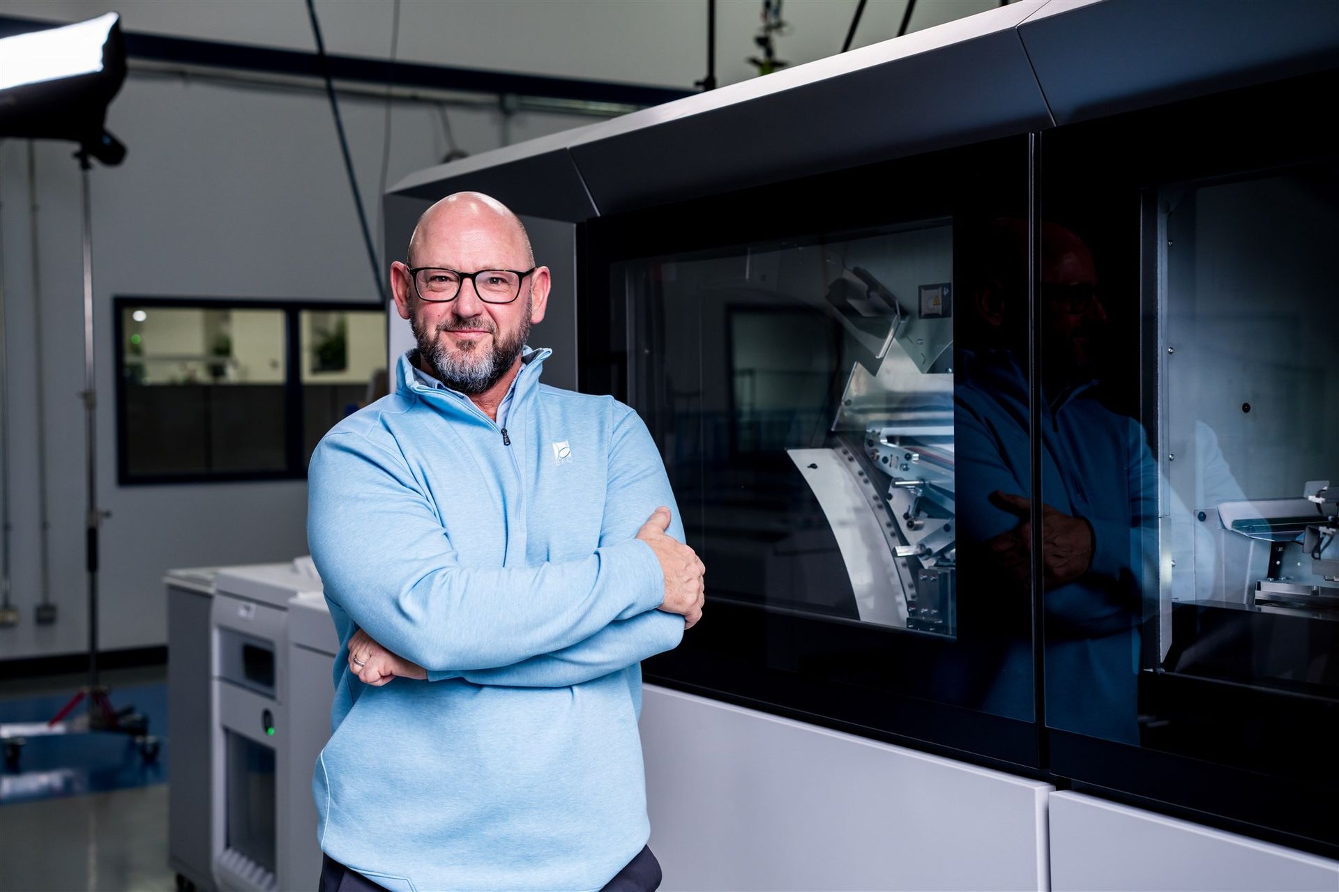 Man in a light blue sweater stands with arms crossed beside industrial equipment in a lab or workshop