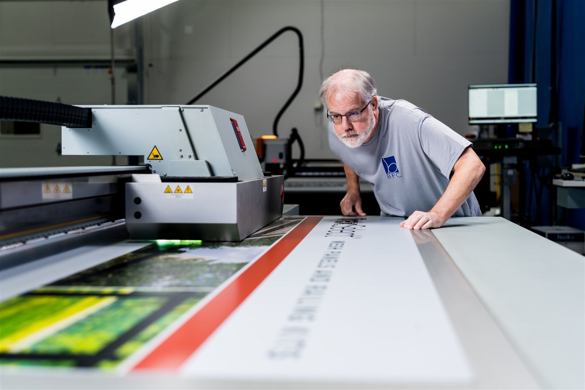 Person inspecting a large-format printer in a print shop, leaning over a sheet of paper