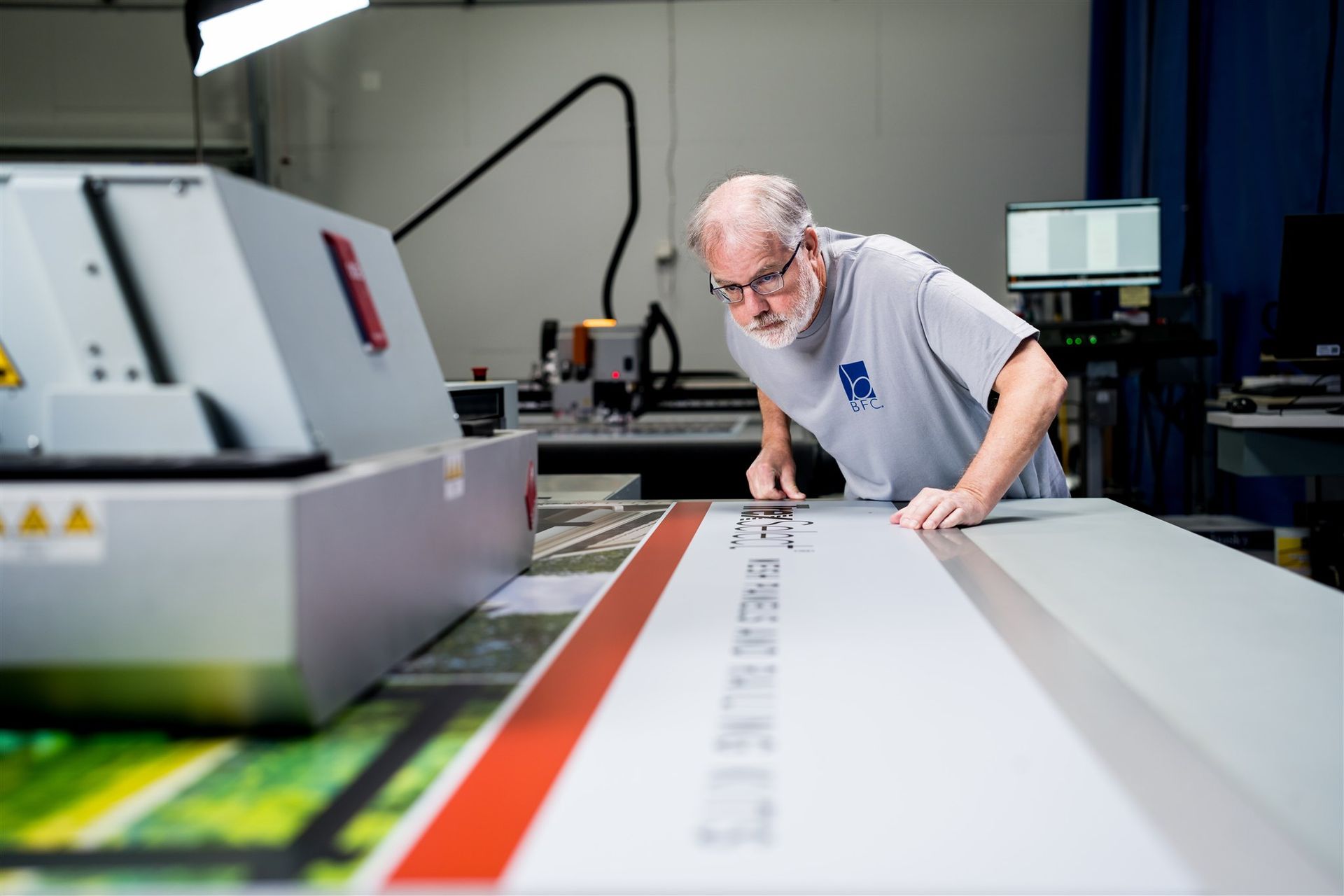 Man inspecting a large-format printer in a print shop, leaning over a long sheet of paper
