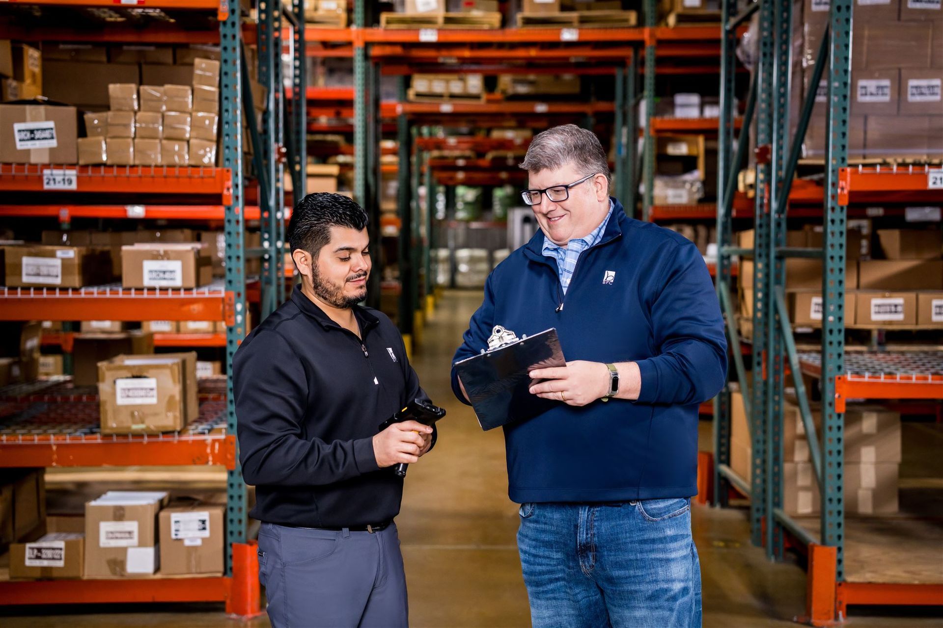 Two warehouse workers stand among shelves of boxed inventory, discussing notes on a clipboard.