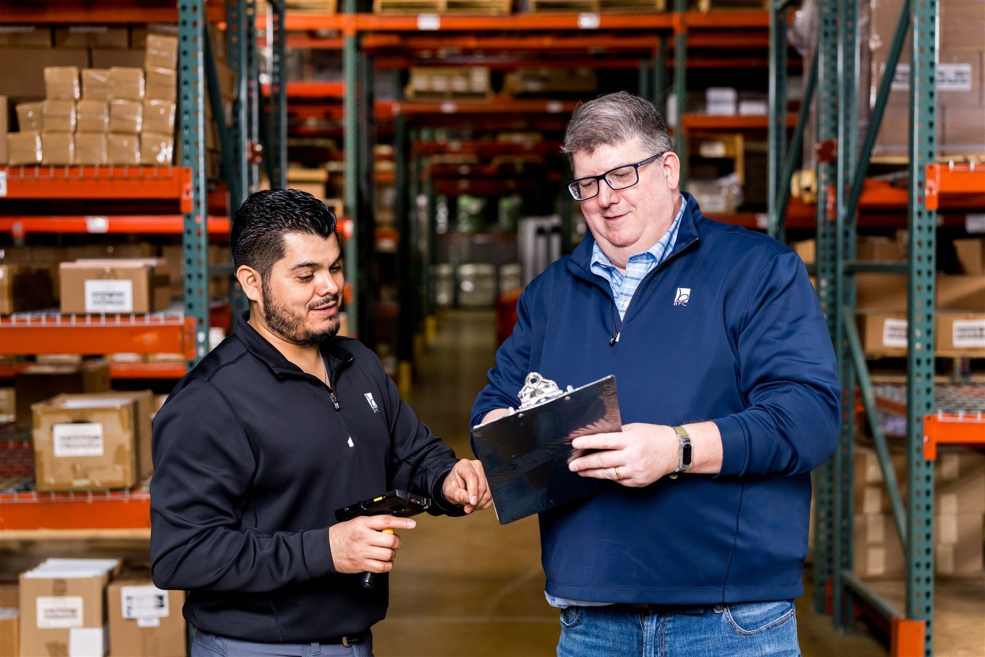 Two coworkers reviewing paperwork in a warehouse aisle with stacked boxes on shelves