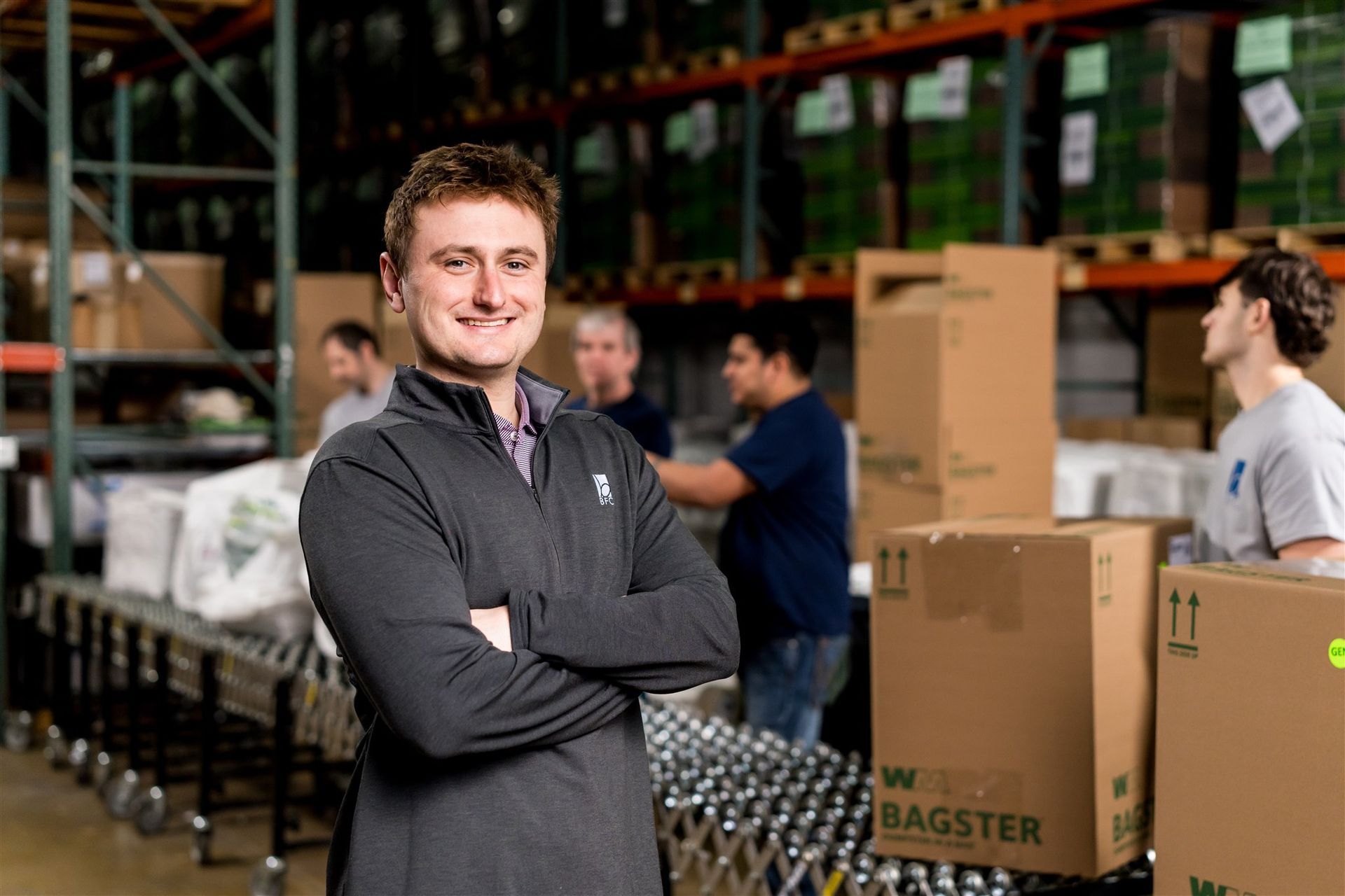 Person standing with arms crossed in a warehouse aisle beside stacked cardboard boxes
