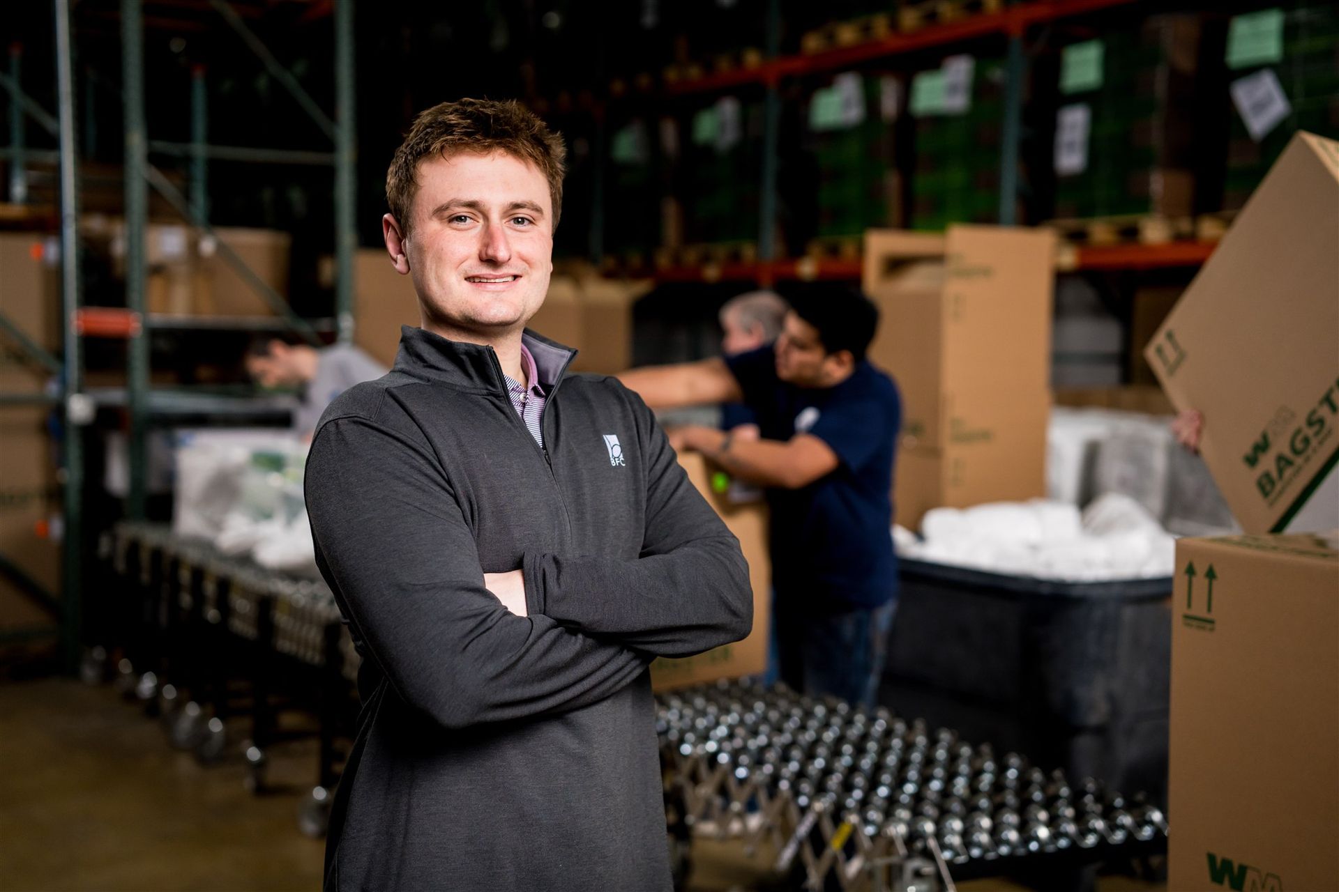 Man standing with arms crossed in a warehouse, with stacked boxes and bottles in the background.