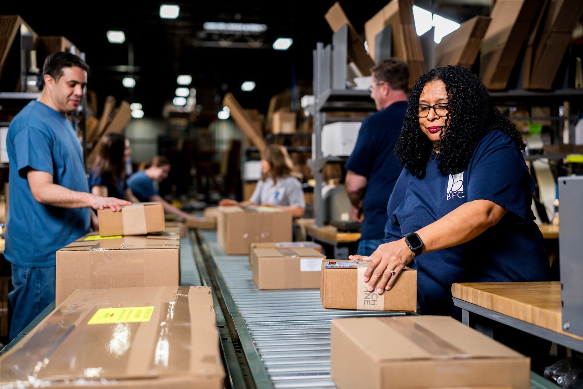 Workers sort packages on a warehouse conveyor belt amid stacks of cardboard boxes.