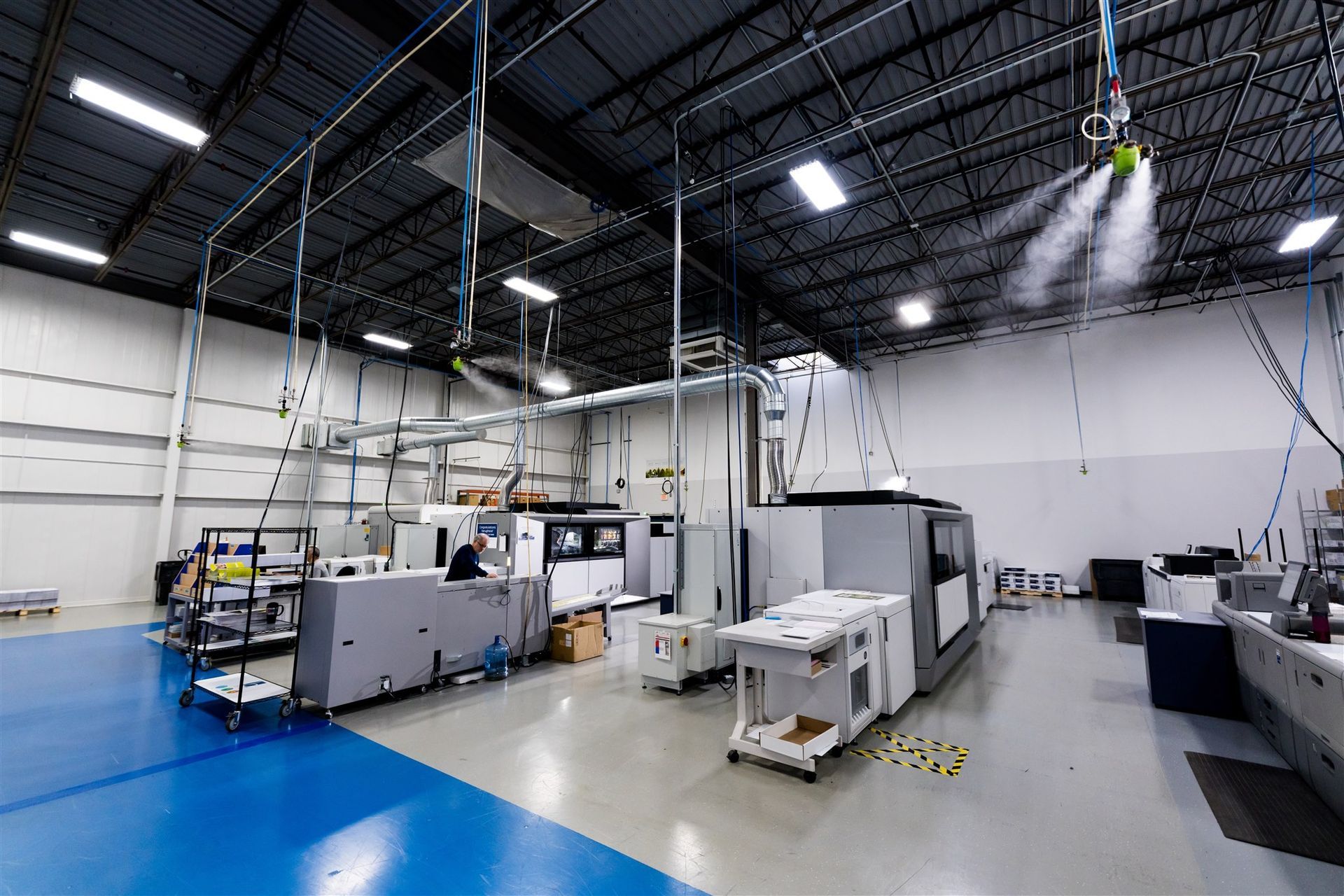 Large industrial lab with printing/machining equipment, blue floor, and high black ceiling on a spacious factory floor