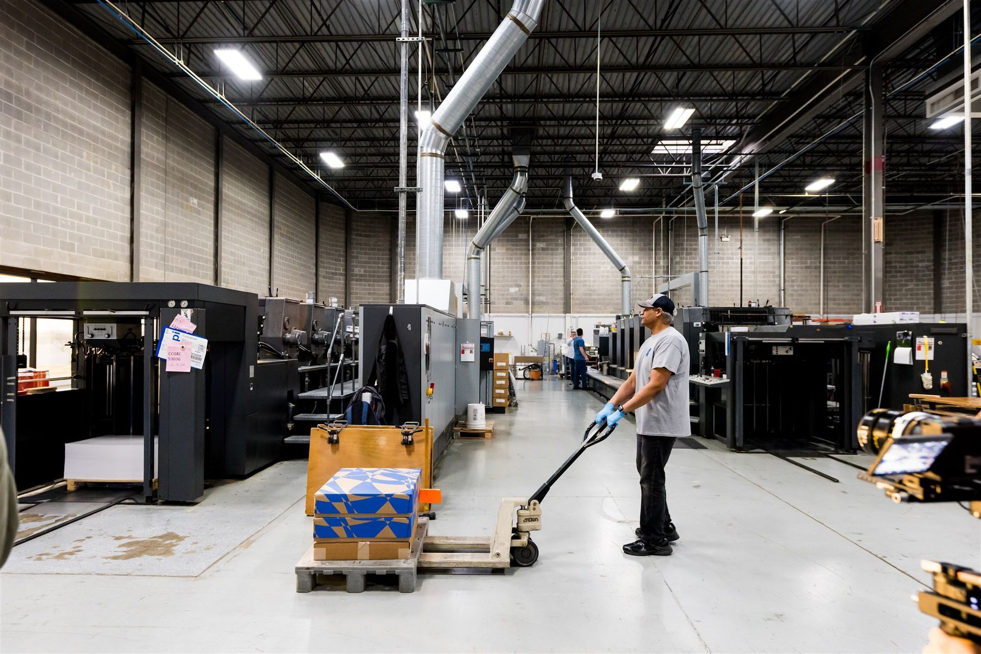 Man in a warehouse aisle moving a pallet jack beside stacked boxes and industrial equipment.