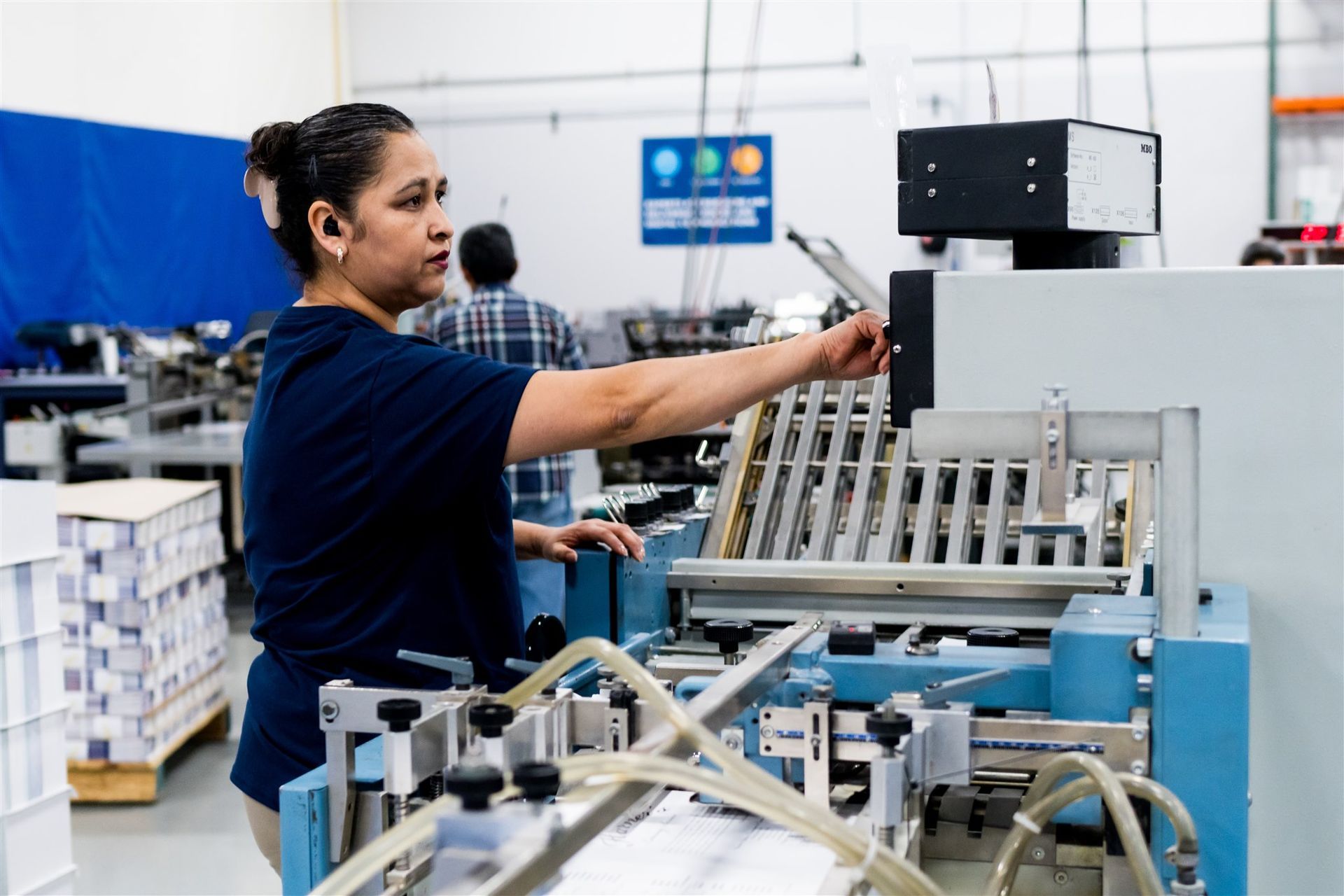 Worker operating machinery in a factory, reaching toward a blue industrial machine with metal parts.