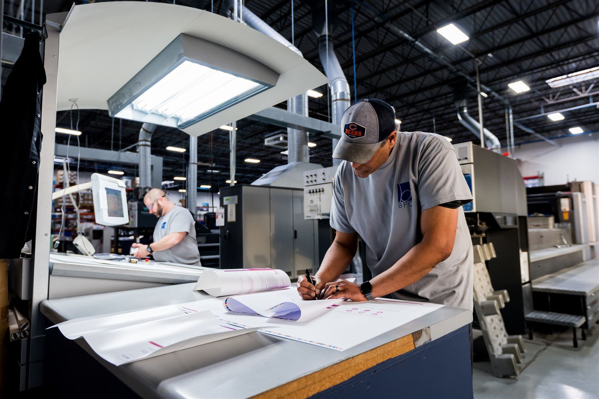 Worker in a print shop reviews papers under bright task lights, with printing equipment in the background.