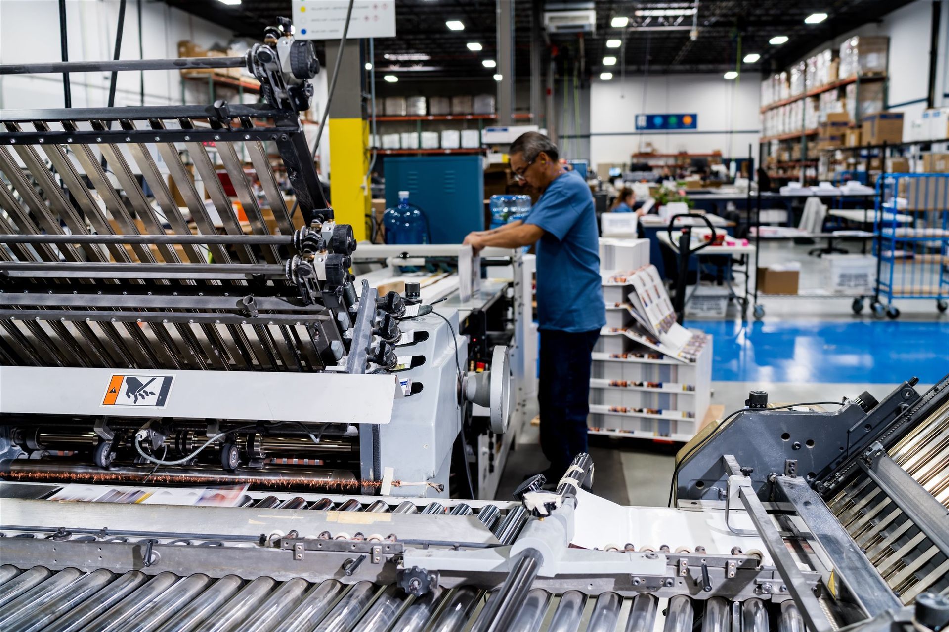 Worker operating machinery in a warehouse assembly line with stacked parts and conveyors