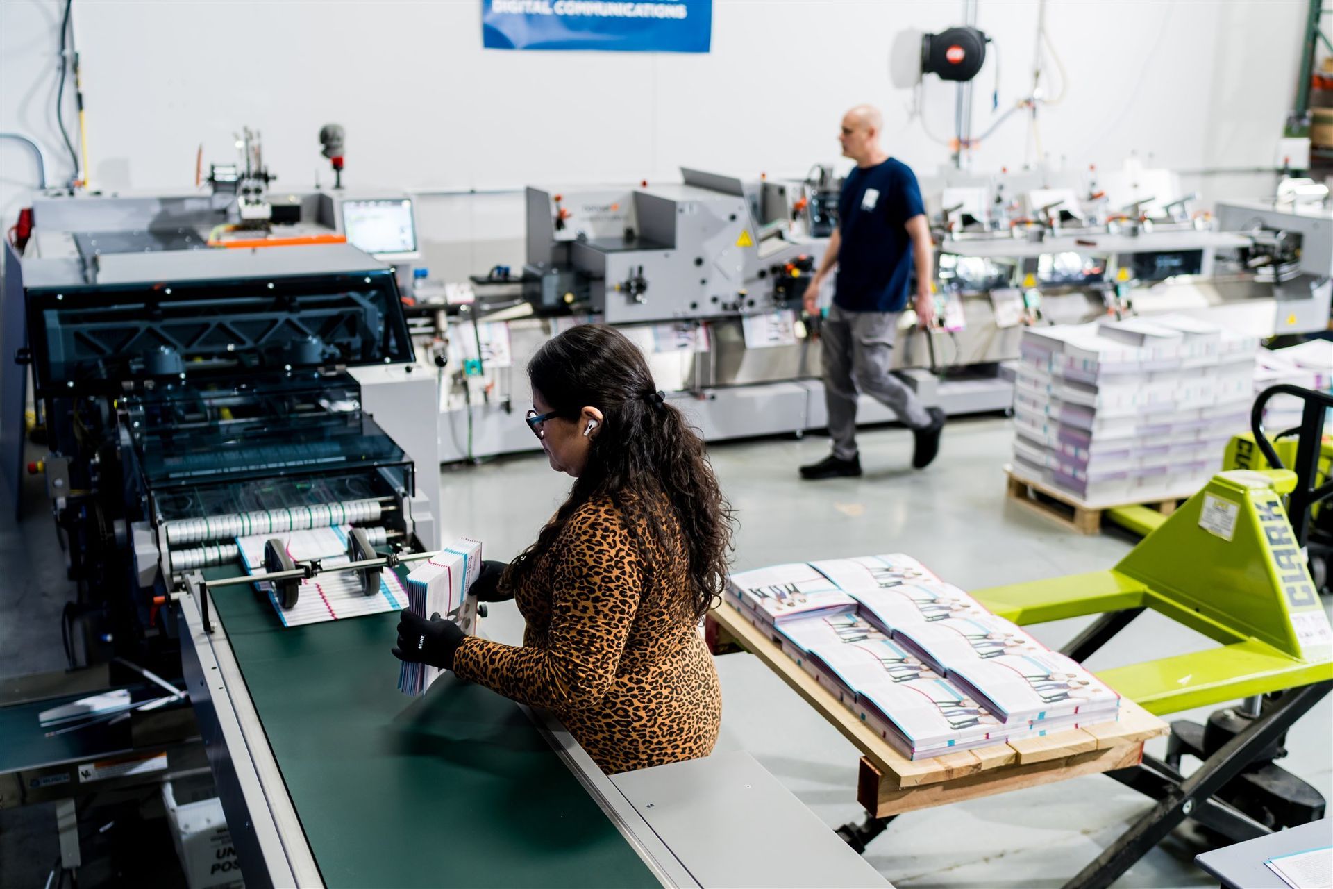 People working at industrial machines in a bright print shop, with stacks of paper and a green pallet jack.