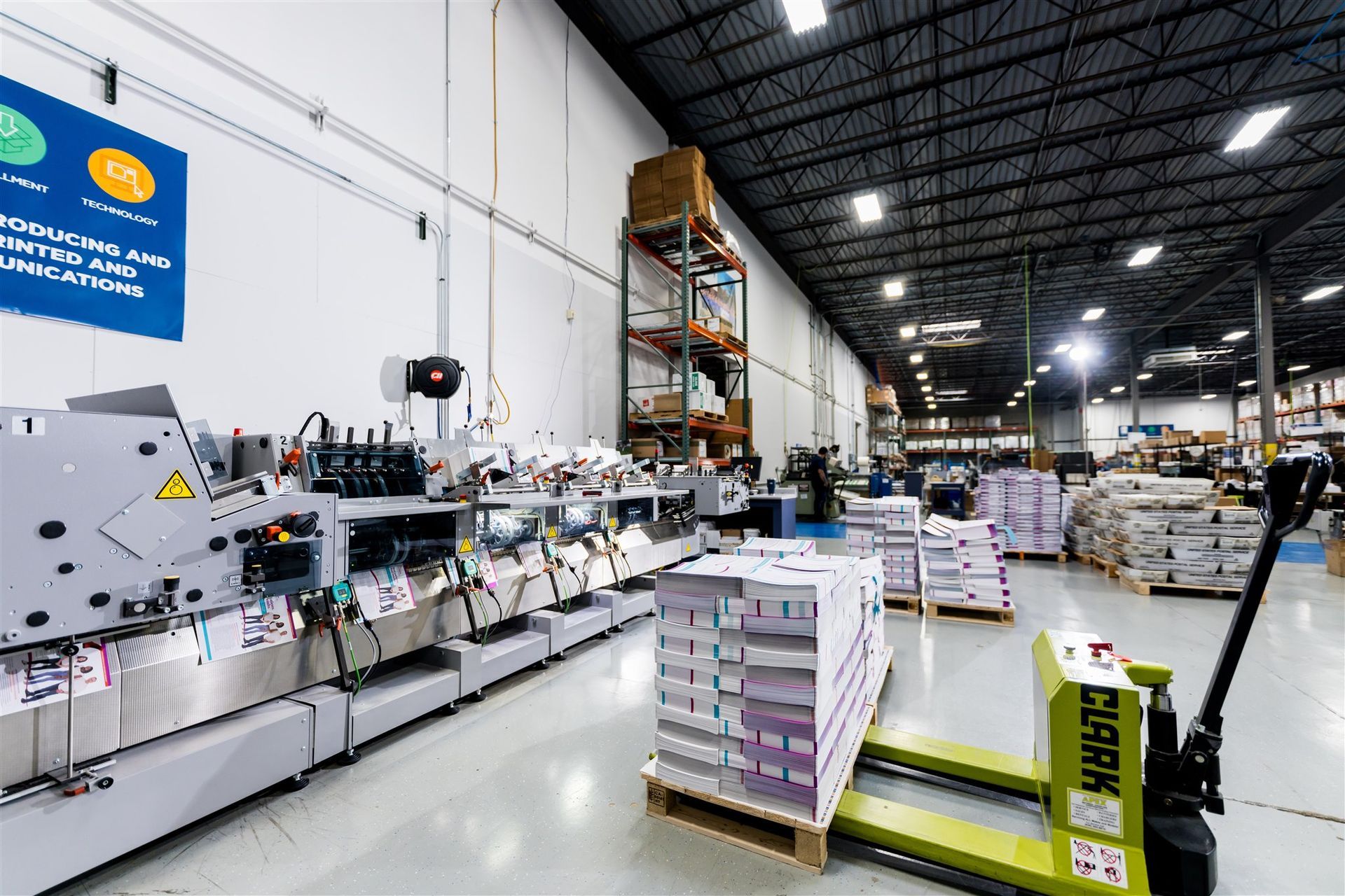 Wide warehouse interior with stacked pallets and a long industrial machine line under bright ceiling lights