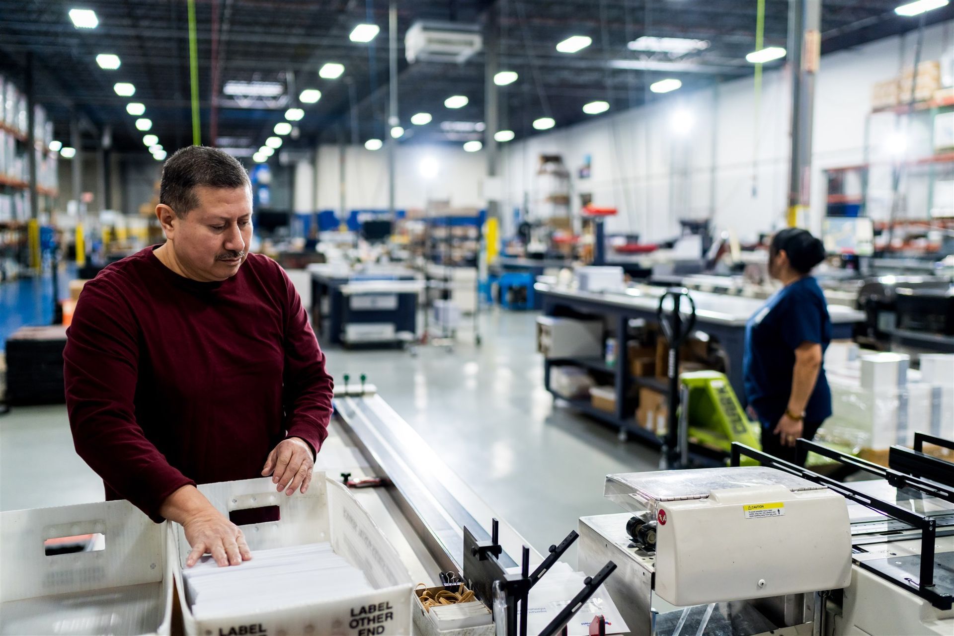 Factory workers operating machinery in a bright industrial workshop, one in a red sweater in the foreground