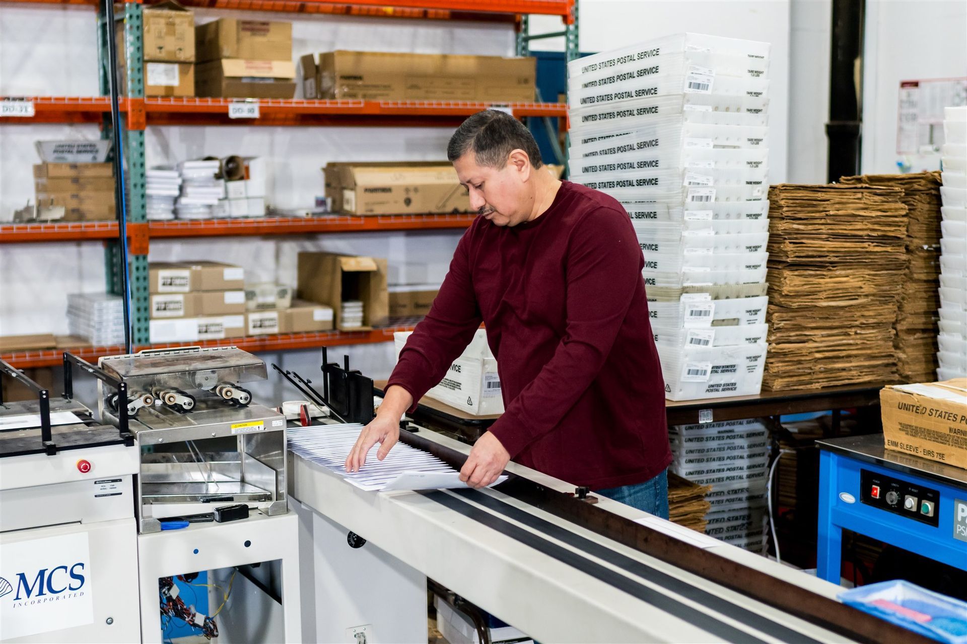 Worker in a maroon shirt using a machine in a warehouse with stacked boxes and shelves