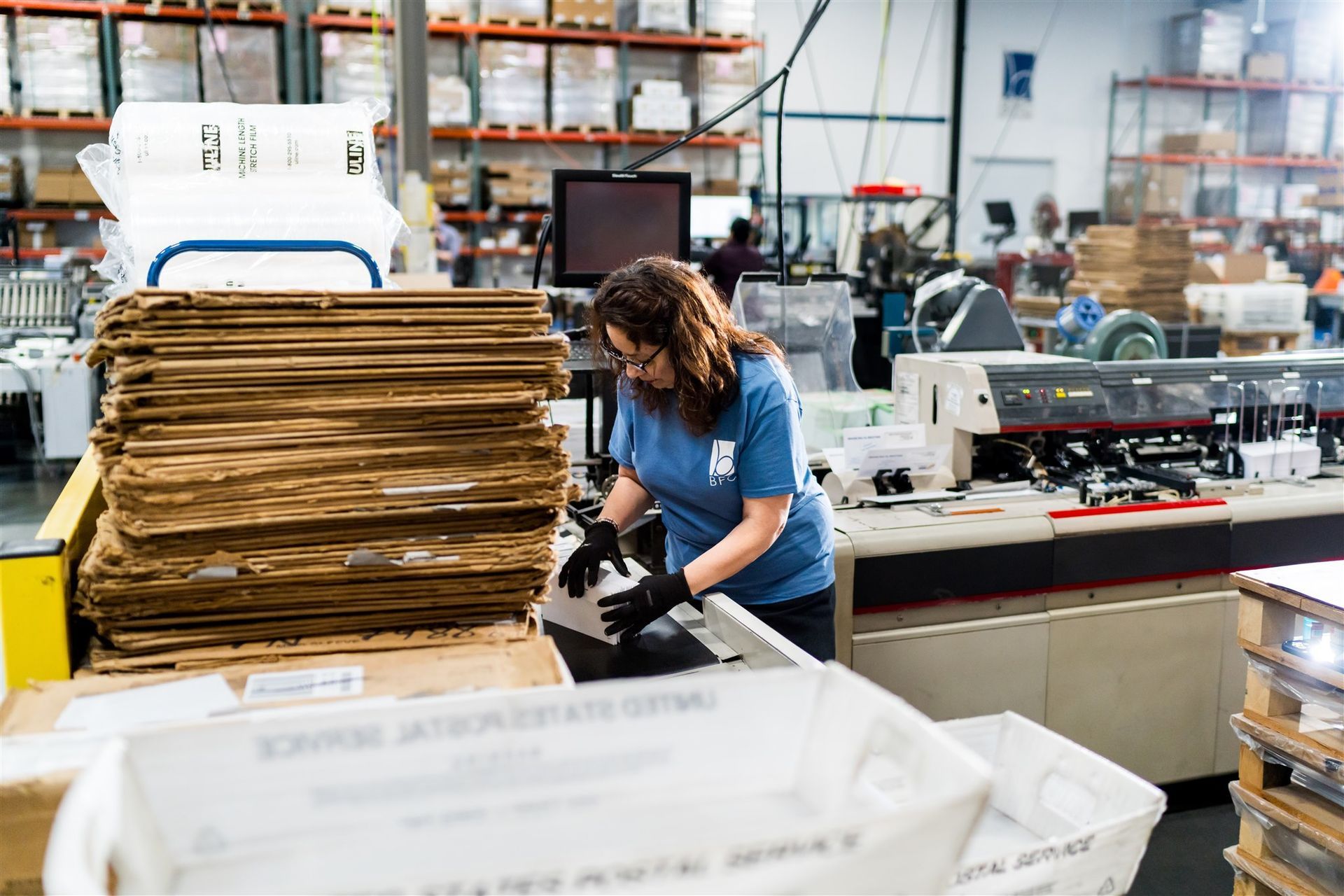 Worker sorting paper goods in a busy warehouse with stacked cardboard bundles and machinery in the background