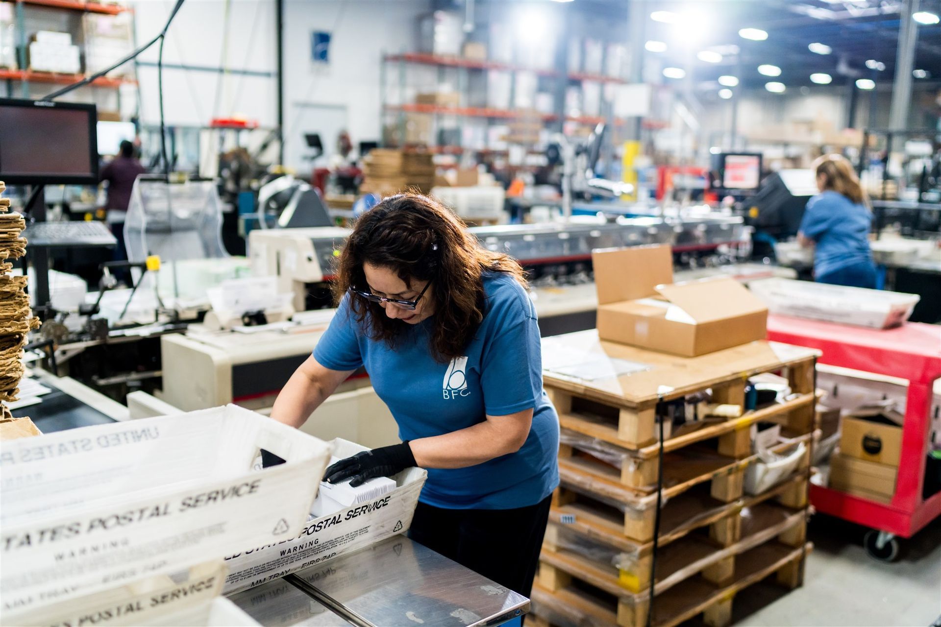 Woman in blue shirt packing items at a warehouse workbench with boxes and pallets nearby