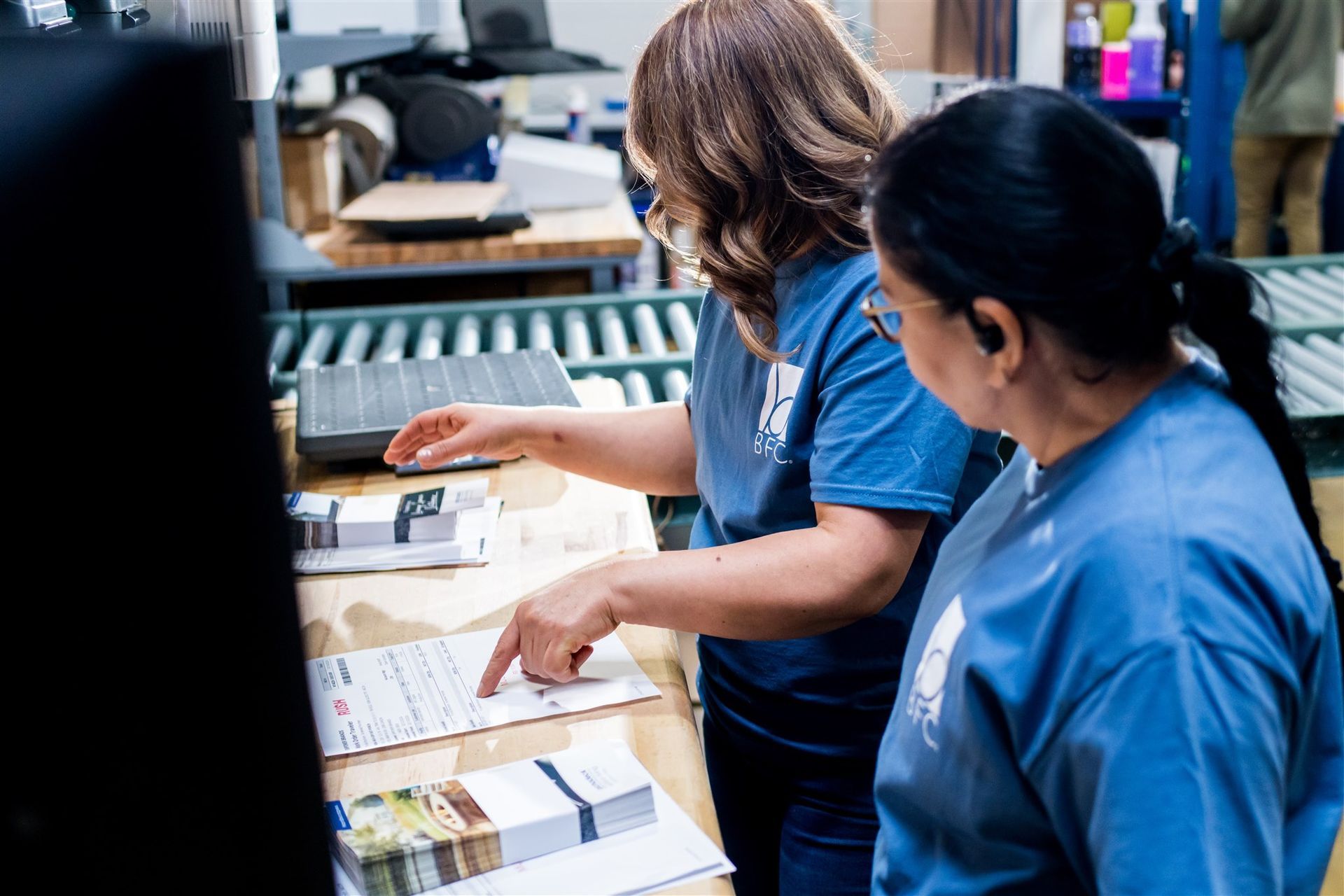 Two workers in blue shirts review papers at a cluttered office desk