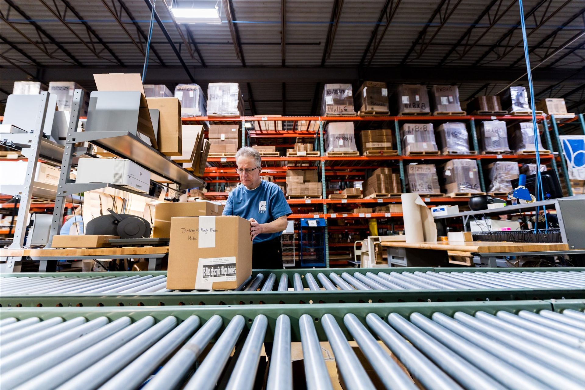 Worker packing a box in a warehouse aisle with shelves and conveyor rollers in the foreground