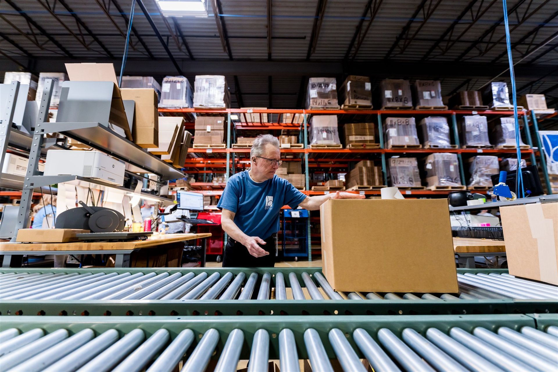 Worker in a warehouse beside a conveyor belt, handling boxes among stacked shelves.