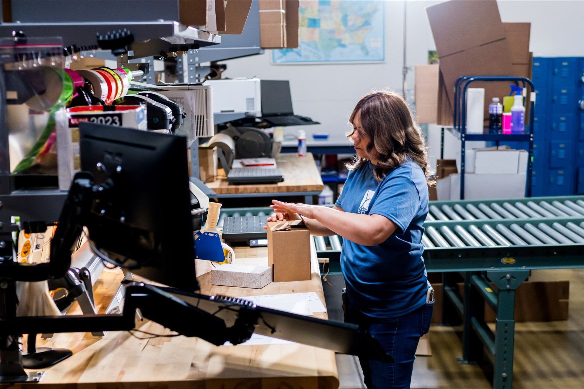 Person working in a cluttered workshop beside a large 3D printer and shelves of supplies.