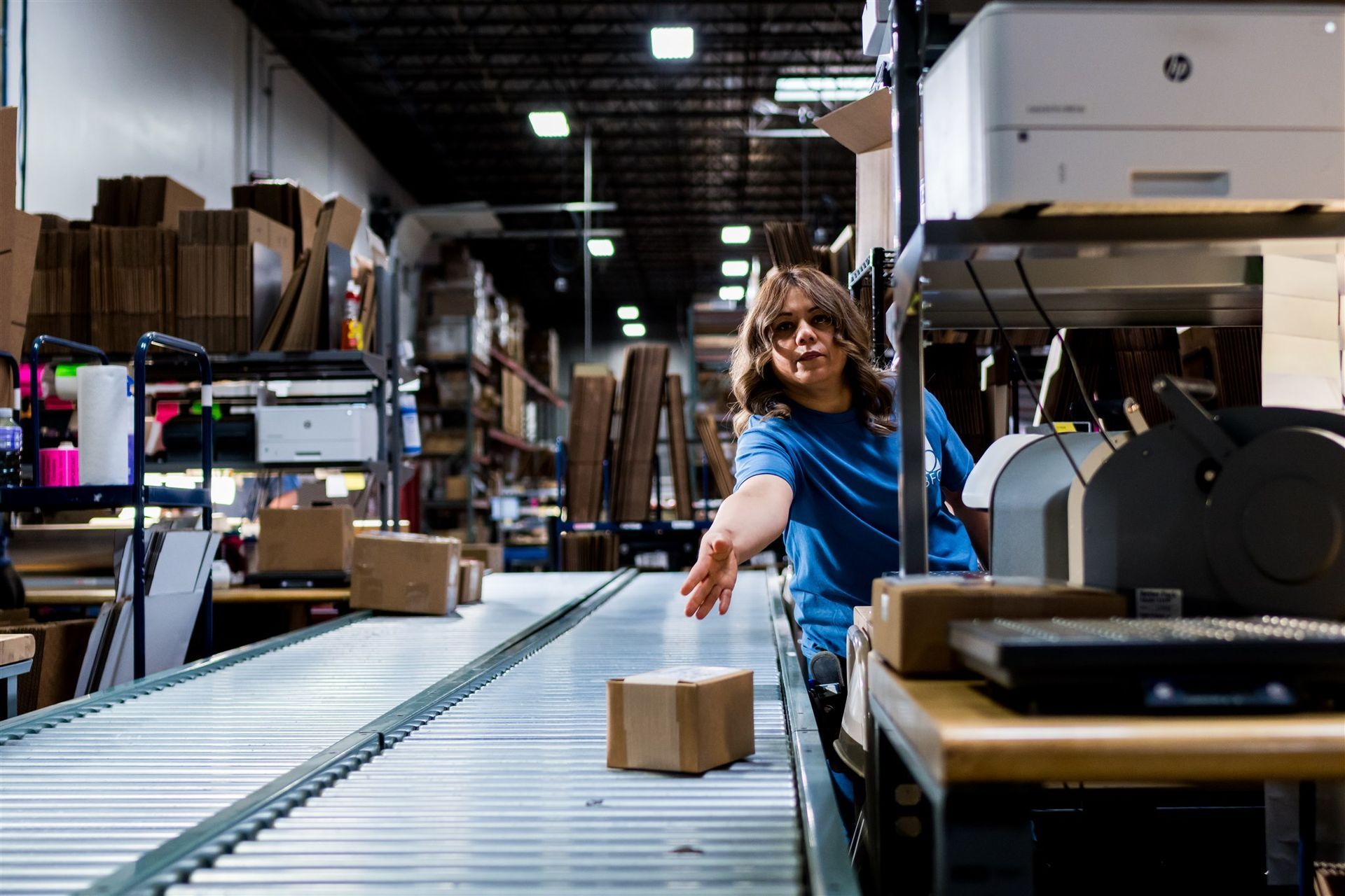 Worker packing a small box on a warehouse conveyor line under industrial lights
